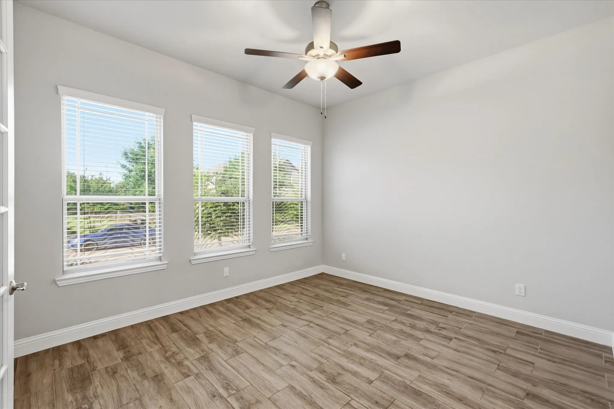 Room featuring three windows with blinds, wood-finish flooring, white baseboards, and a ceiling fan with light fixture