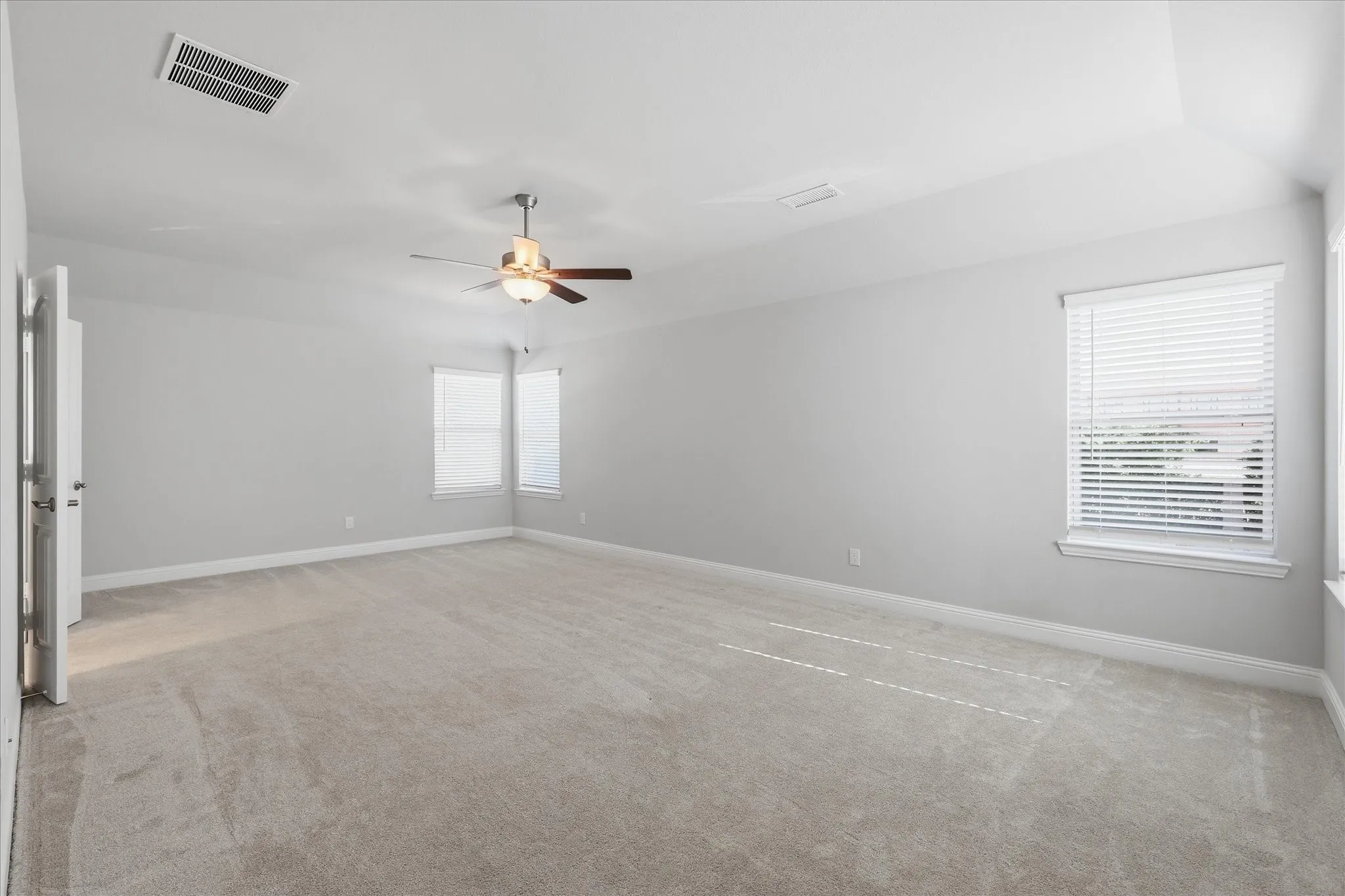 Spacious room featuring light gray carpet, multiple windows with blinds, a ceiling fan with integrated lighting, and a white interior door