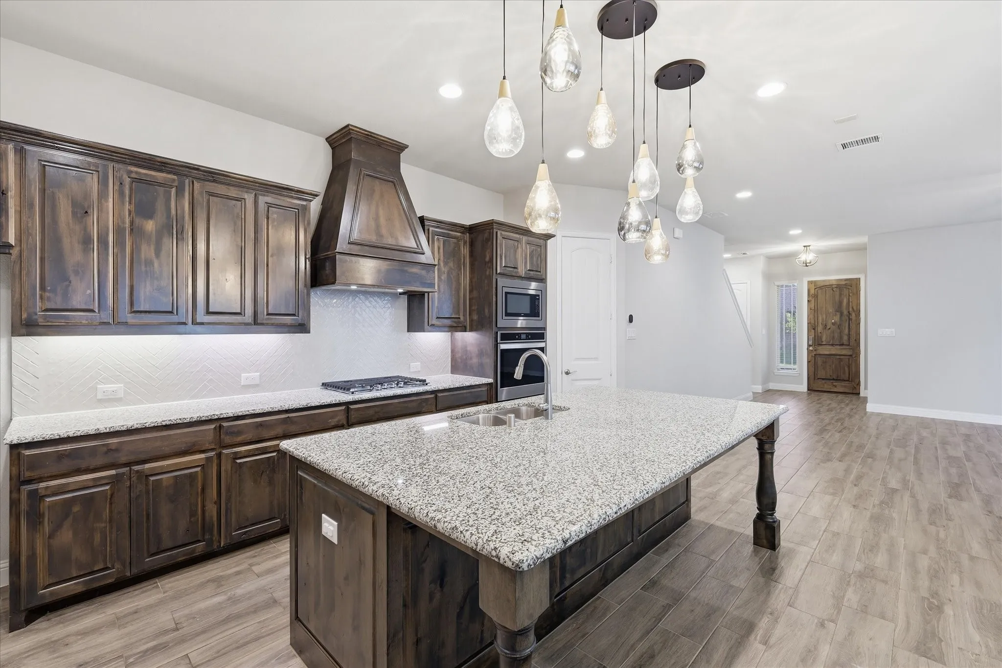 Open-concept kitchen featuring an oversized island with pendant lighting, rich wood cabinetry, light granite countertops, a tiled backsplash, and wood-finish flooring