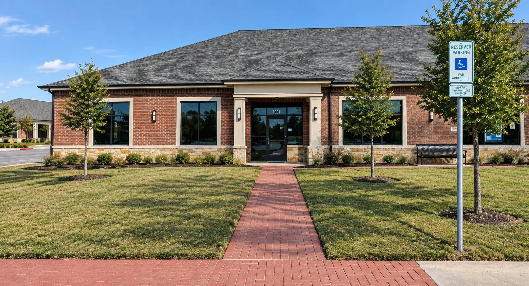 Brick facade building featuring a dark shingle roof, stone foundation, and a red paver walkway