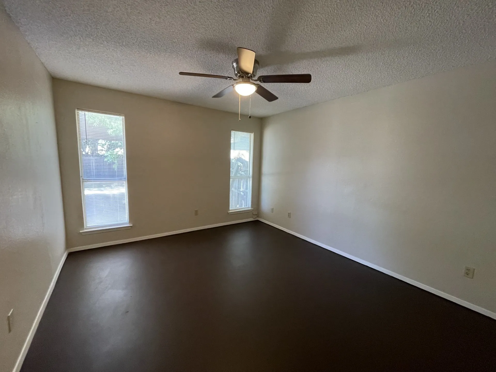 Spacious room featuring dark wood-finish flooring, two large windows, white trim, and a ceiling fan with integrated lighting
