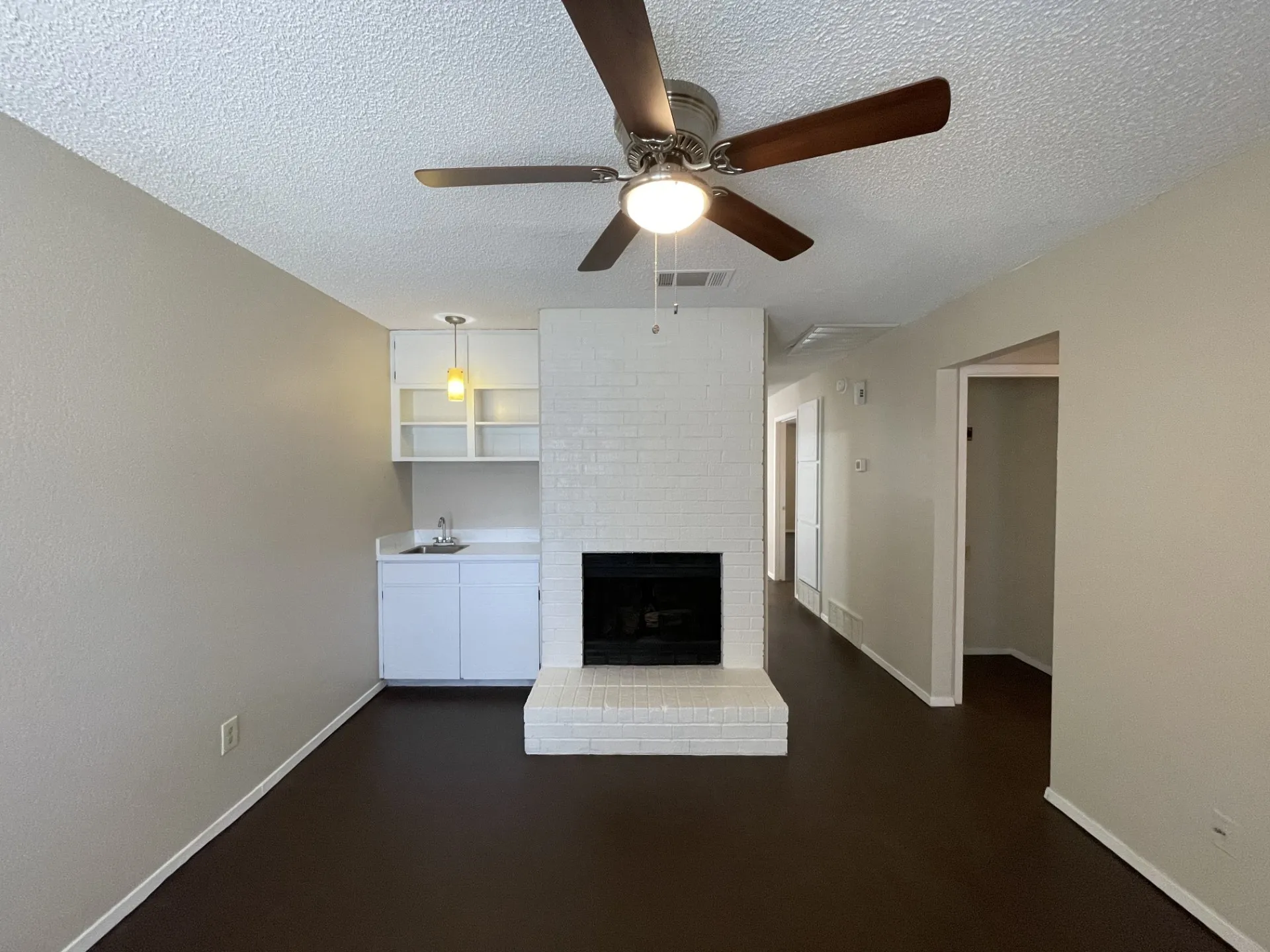 Living space featuring a whitewashed brick fireplace with hearth, a built-in wet bar with a white cabinet and sink, a ceiling fan with light, and dark wood-finish flooring