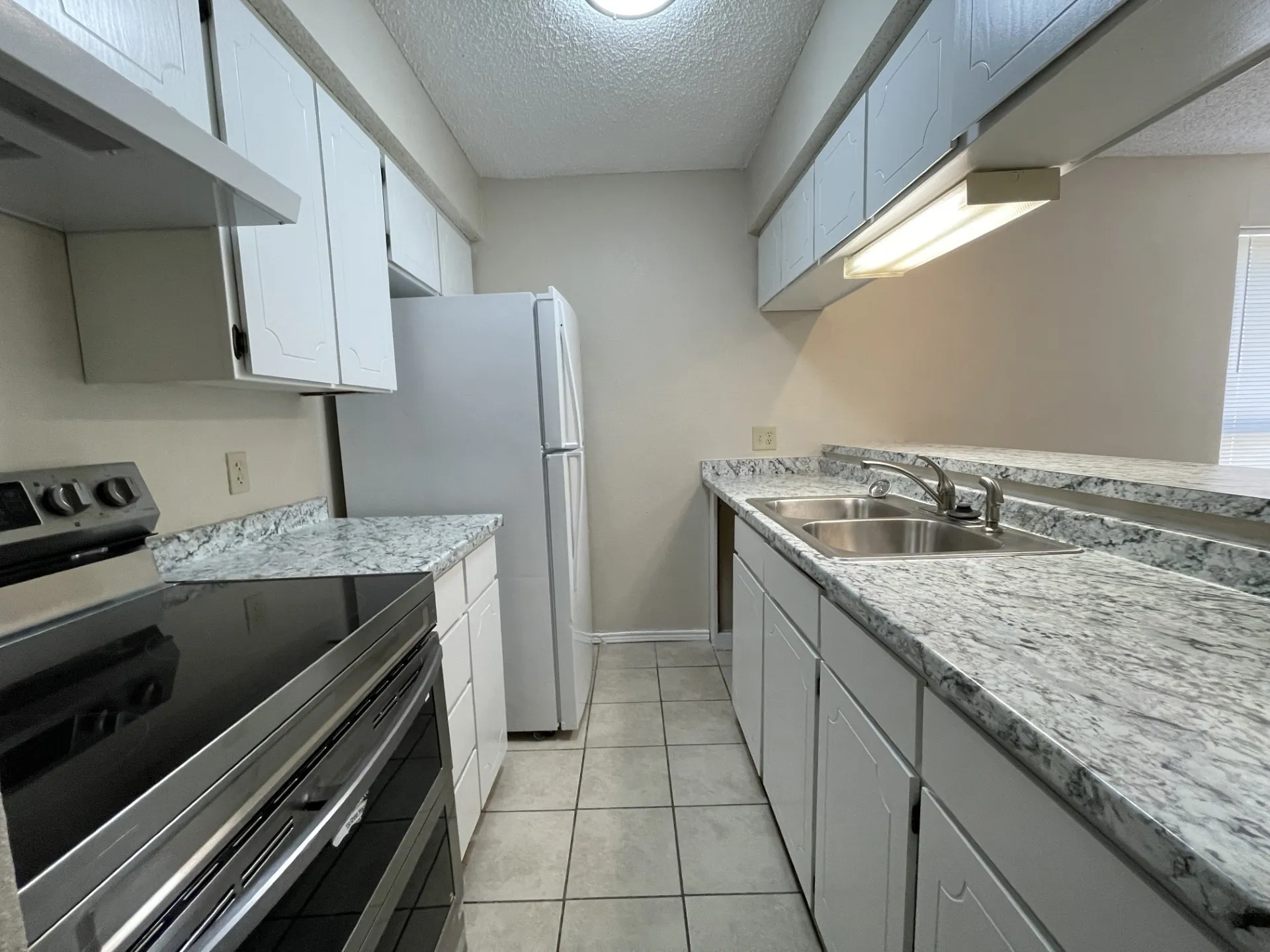 Galley kitchen featuring white cabinetry, gray and white patterned countertops, stainless steel double basin sink, and tile flooring
