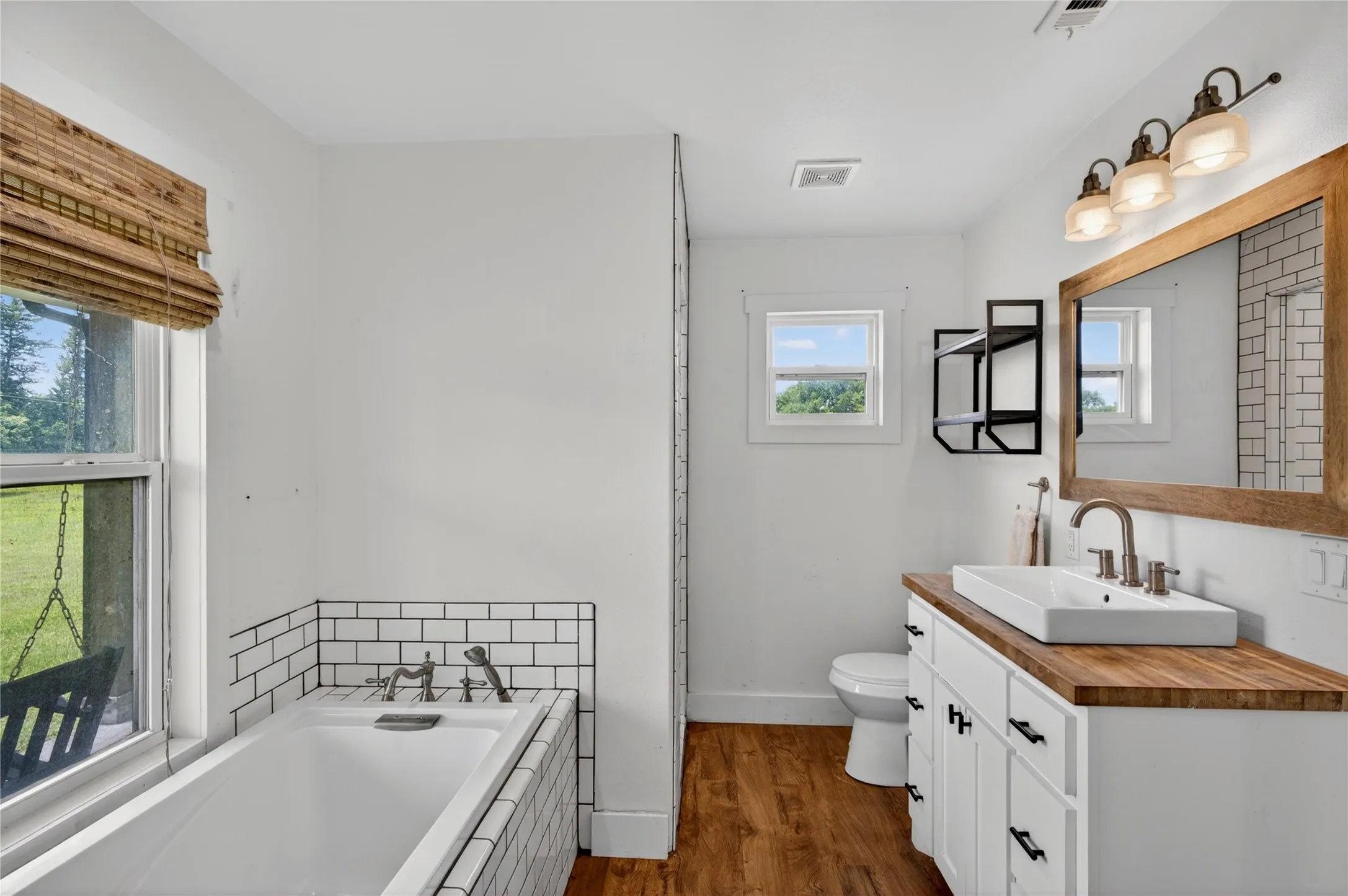 Bathroom featuring a vessel sink vanity with a wood-finish countertop, white cabinetry, a large wood-framed mirror, and a soaking tub with subway tile surround
