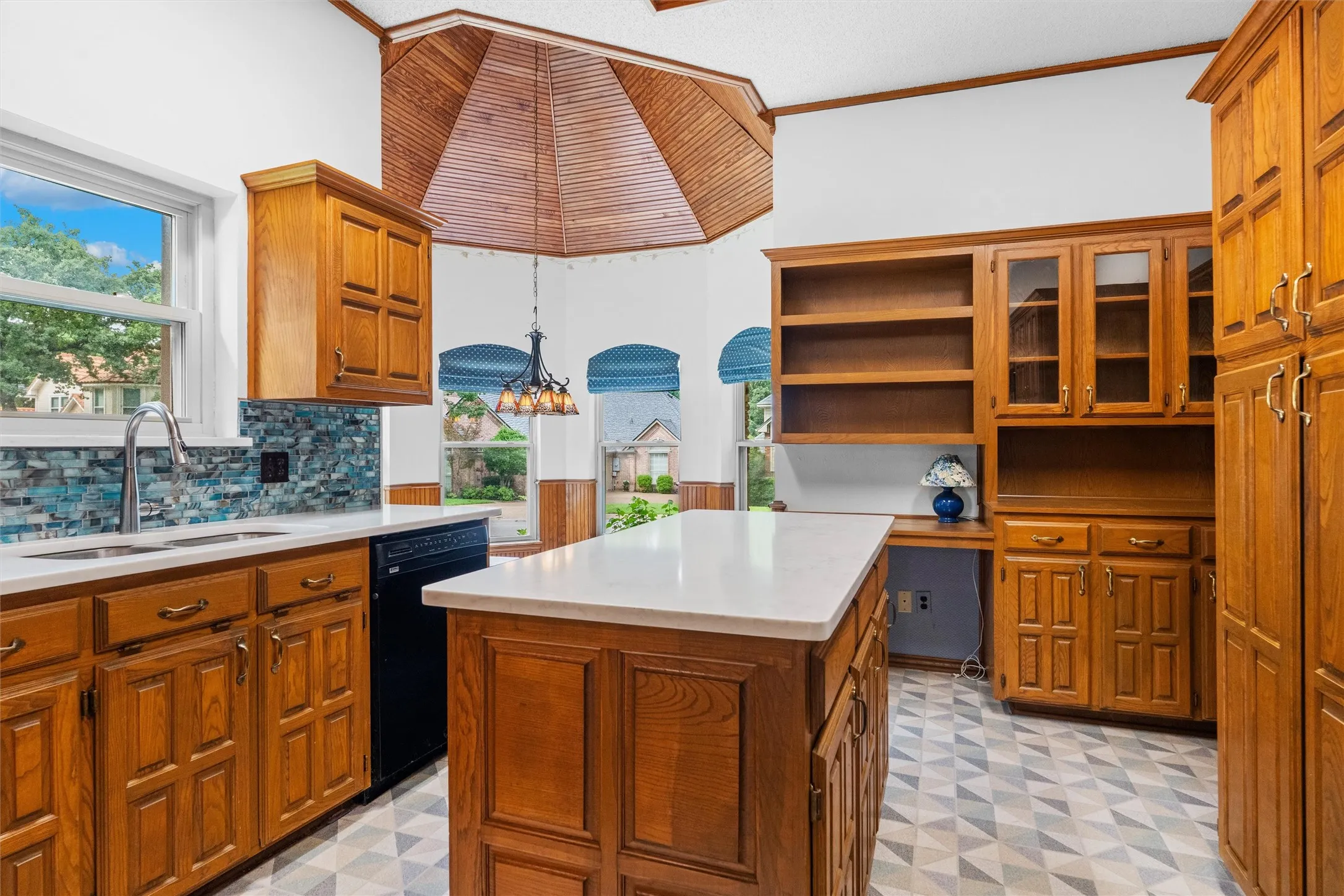 Kitchen featuring a central island with white countertop, extensive oak cabinetry, a built-in desk area, and a vaulted wood-paneled ceiling