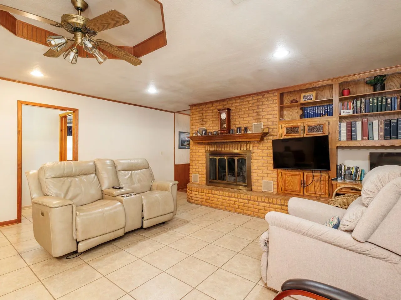 Living area featuring a brick fireplace with a wood mantle, built-in shelving, tile flooring, and a ceiling fan with light fixture
