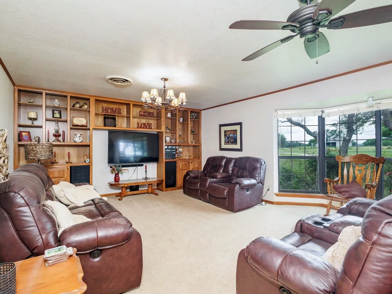 Living room featuring a large bay window, light neutral carpeting, a ceiling fan, and a built-in entertainment center with extensive wood shelving and cabinetry