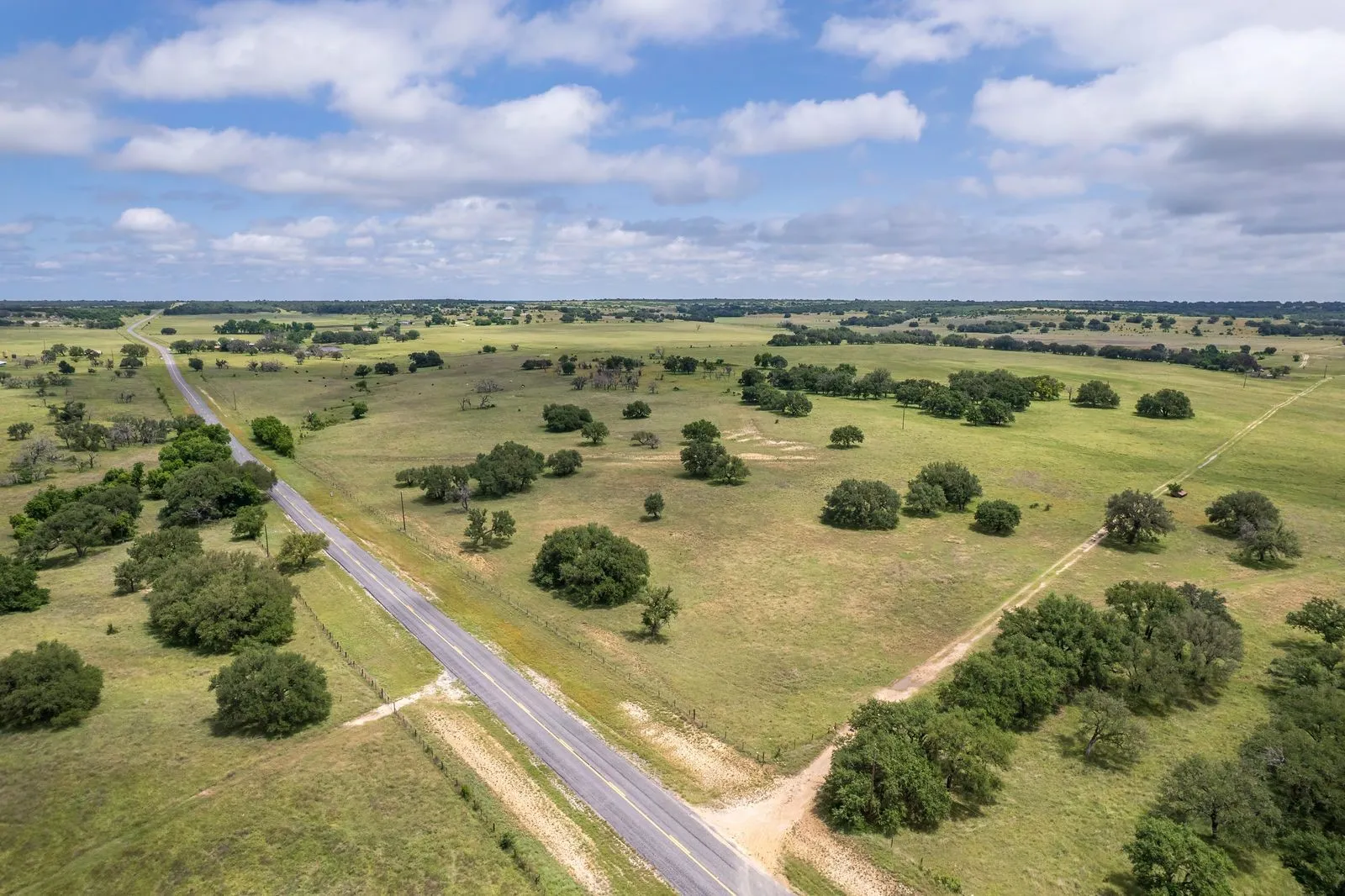 Expansive land parcel featuring a paved road frontage, established tree clusters, open grassy fields, and an unpaved access road