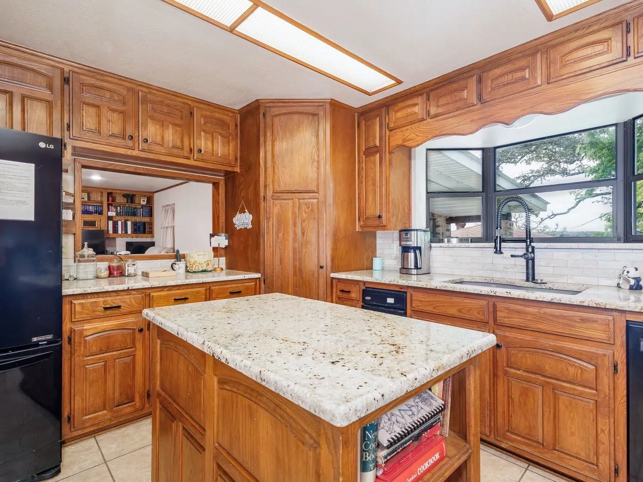 Kitchen featuring a central island with a granite countertop, wood-finish cabinetry, a white subway tile backsplash, a built-in sink with a matte black faucet, and a pass-through window