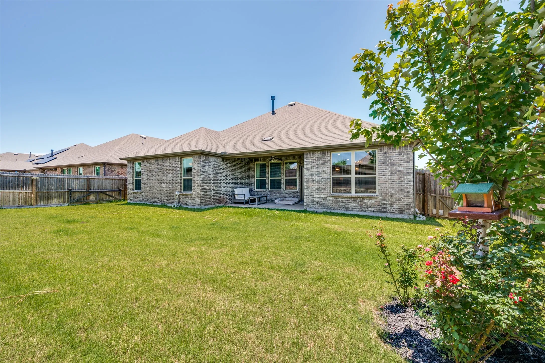Expansive backyard featuring a manicured lawn, brick exterior, concrete patio, perimeter fencing, and mature landscaping
