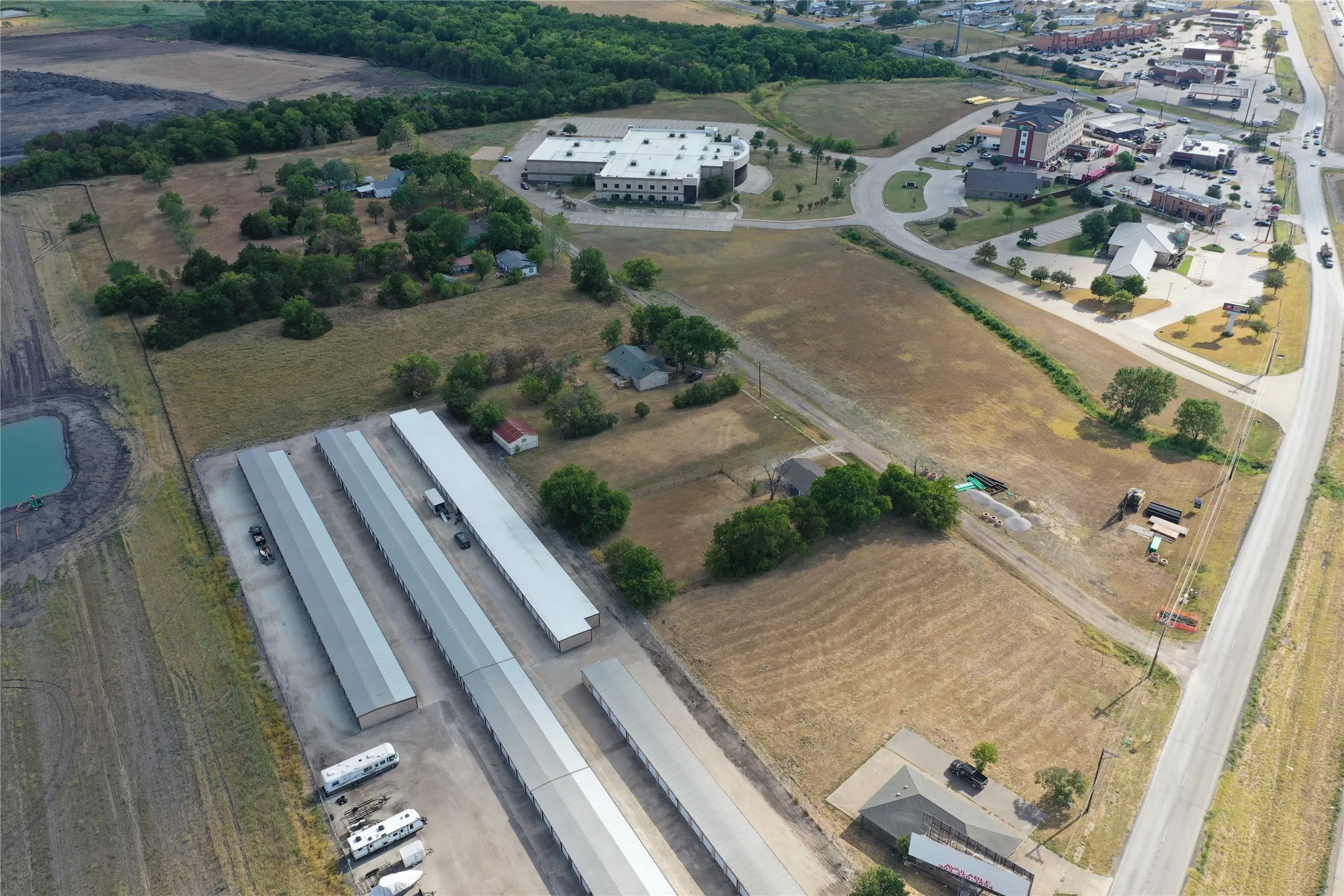 Aerial perspective of an expansive land parcel featuring existing self-storage facilities, a commercial building with visible parking, and a prominent roadway frontage