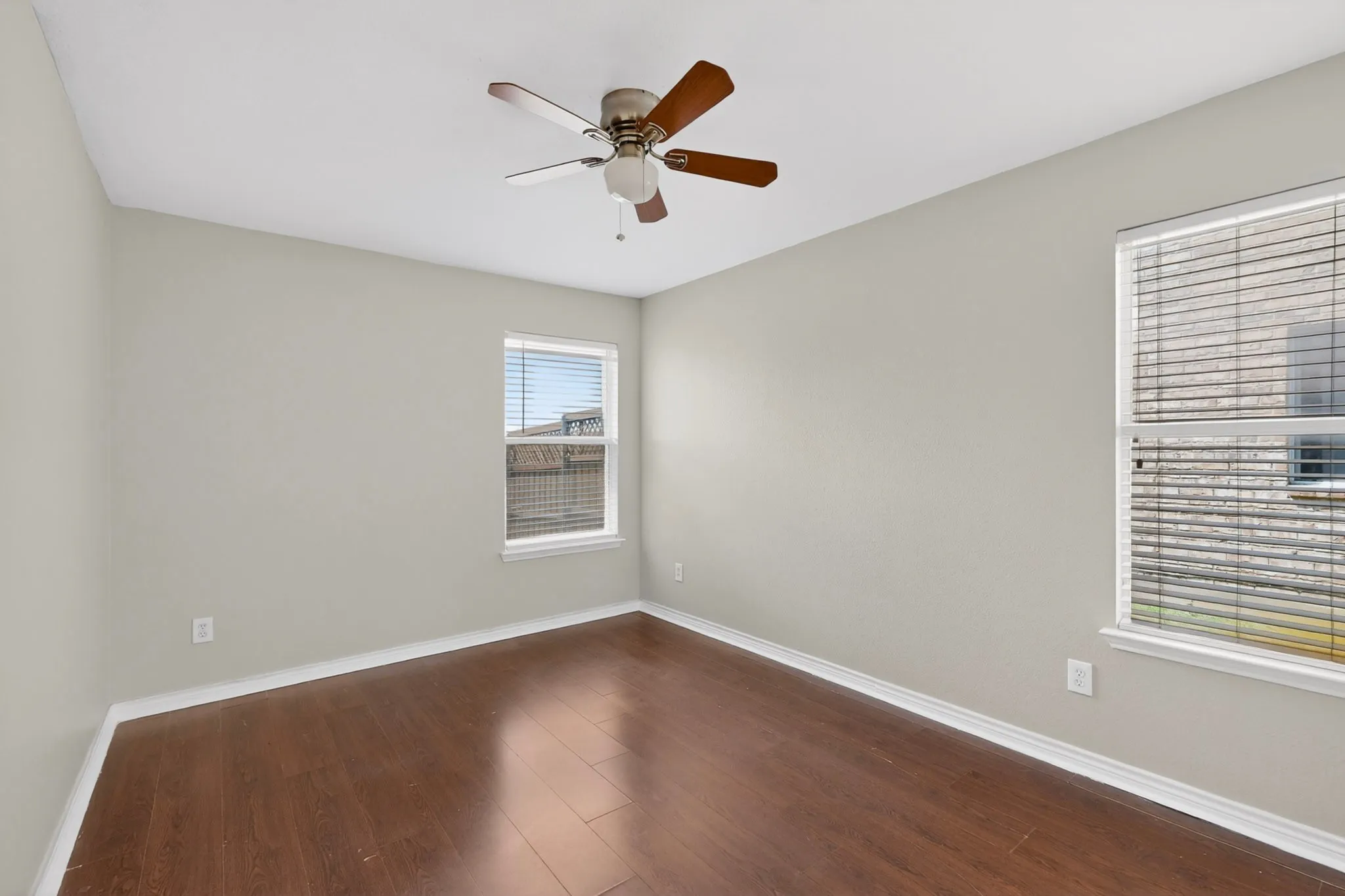 Bright room featuring wood-finish flooring, two large windows with blinds, a ceiling fan with light fixture, and neutral painted walls with white trim