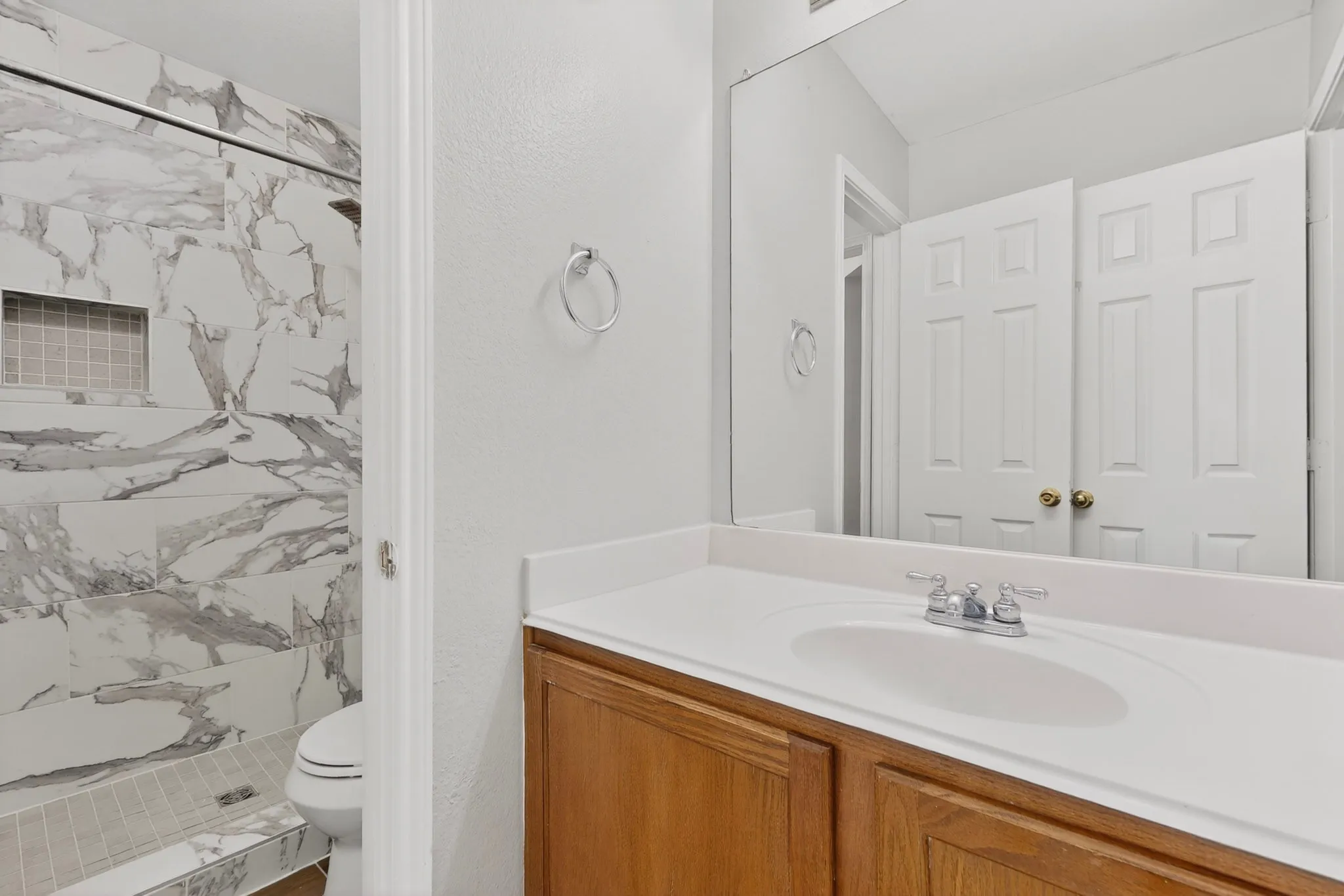 Bathroom featuring a wood-finish vanity with an integrated sink, a large wall-mounted mirror, and a shower with marble-look tile surround and a recessed niche