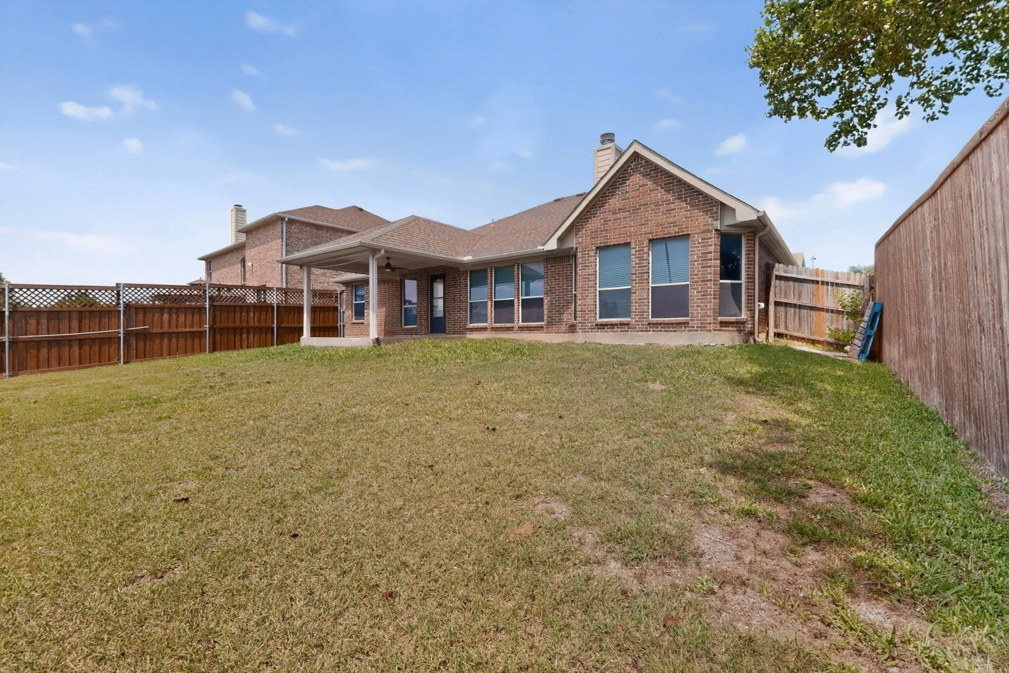 Expansive backyard featuring a covered patio, brick exterior, multiple windows, and a wood privacy fence