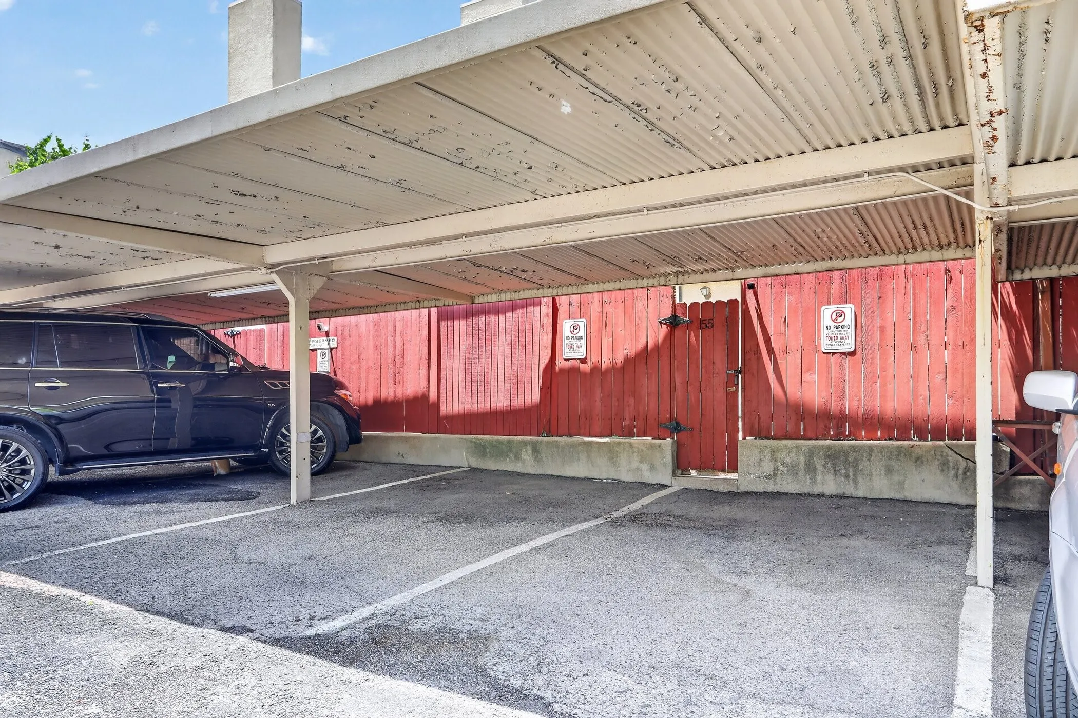 Carport with designated parking spaces, corrugated metal roofing, asphalt paving, and a stained wood fence