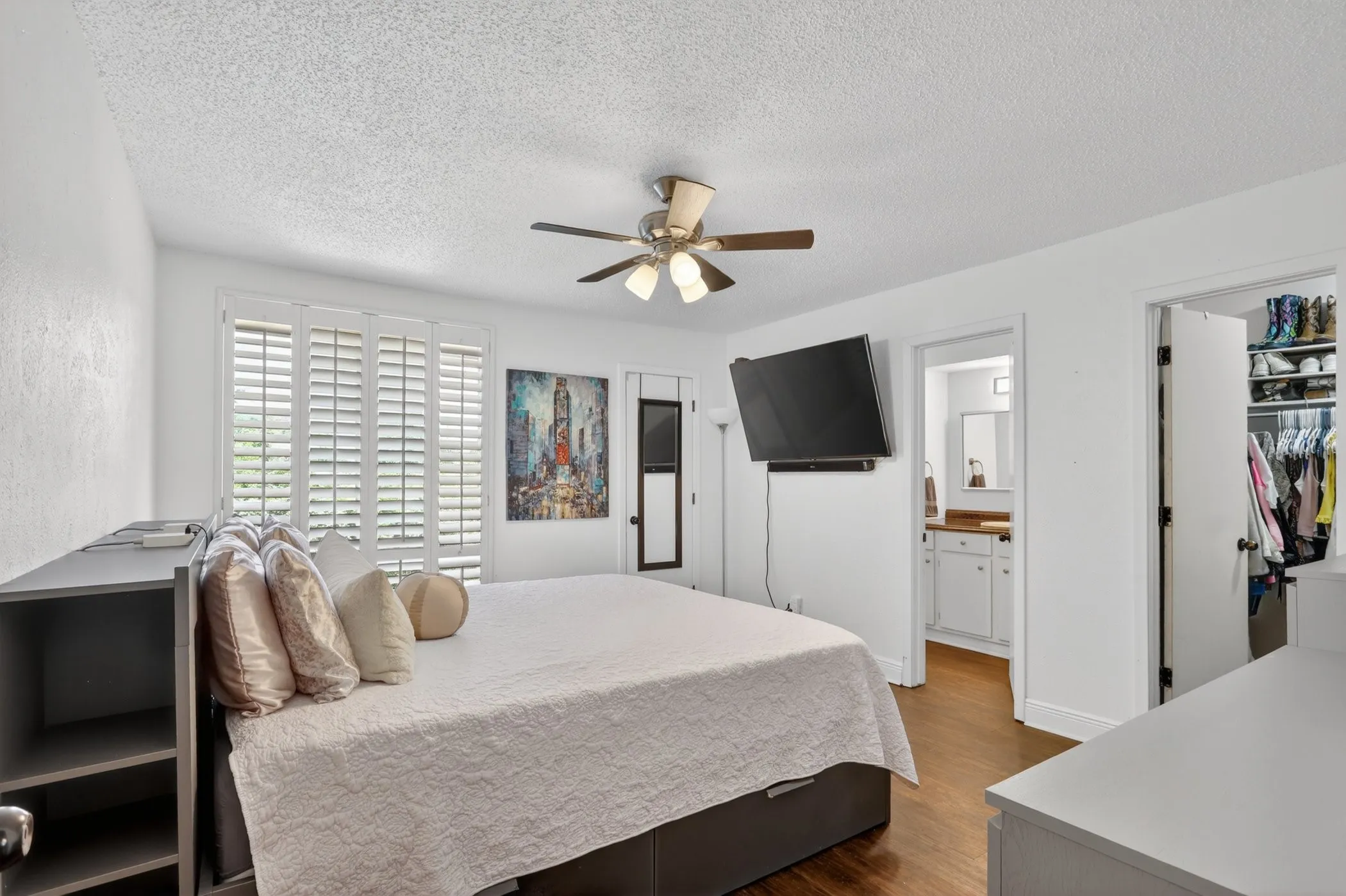 Bedroom featuring wood-finish flooring, built-in shelving, a ceiling fan with integrated lighting, window shutters, and an attached bathroom with a vanity