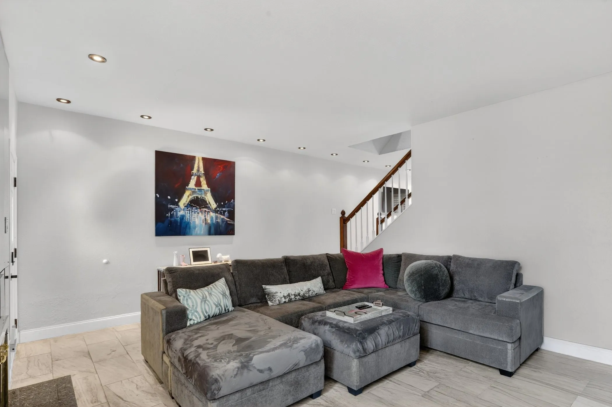 Recessed ceiling lighting illuminates a living area featuring light-toned flooring, white walls, and a visible staircase with wooden handrails and white balusters