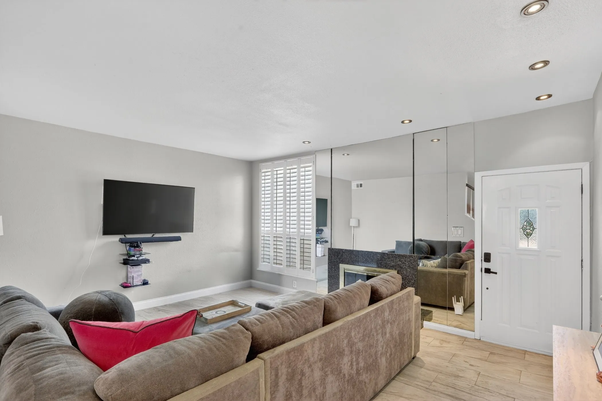 Living area with tile-finish flooring, recessed lighting, white interior door with decorative glass inlay, full-wall mirrored accent, and plantation shutters