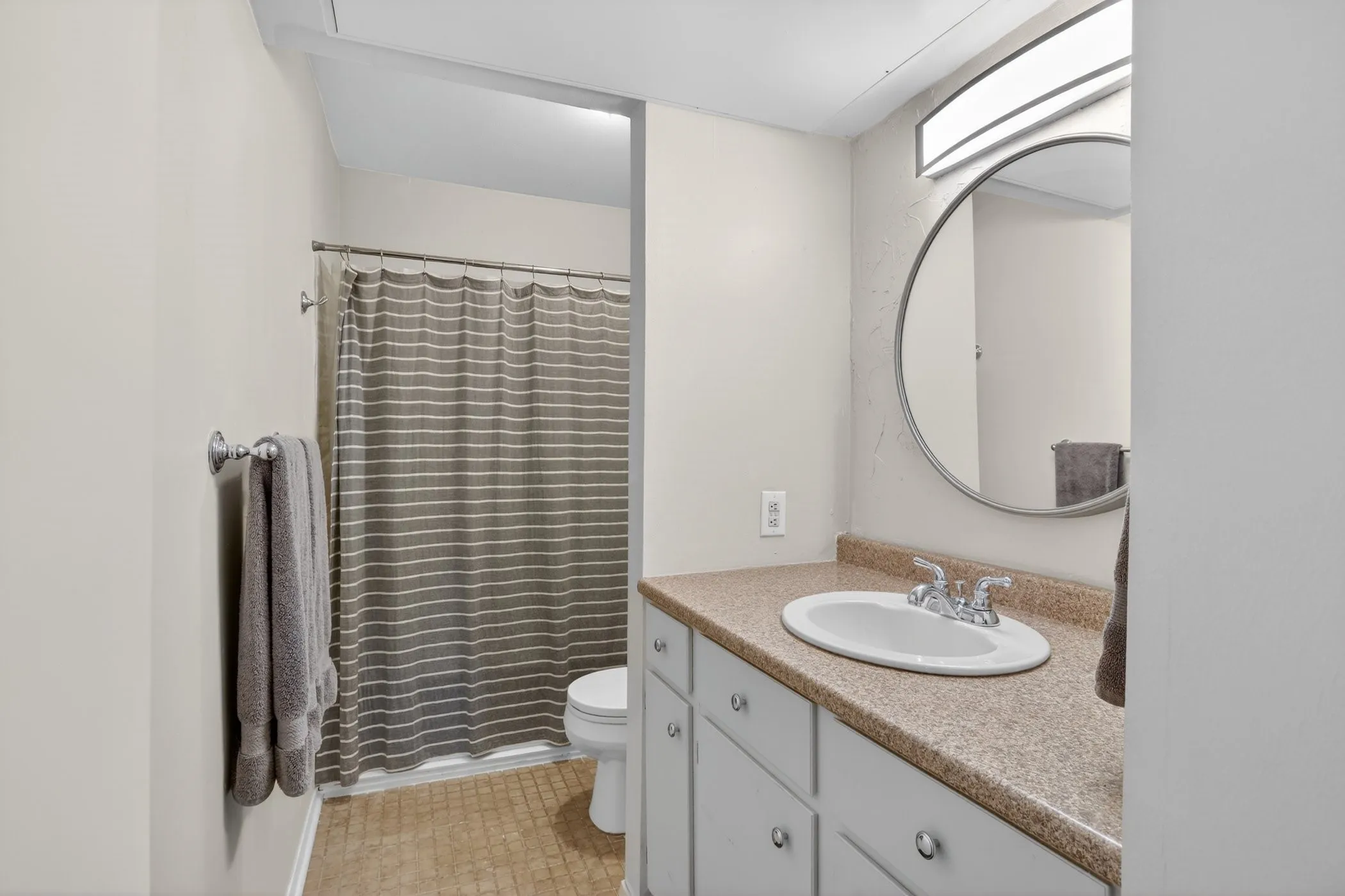 Bathroom featuring a white vanity with a tan countertop, an oval undermount sink, and a circular wall-mounted mirror
