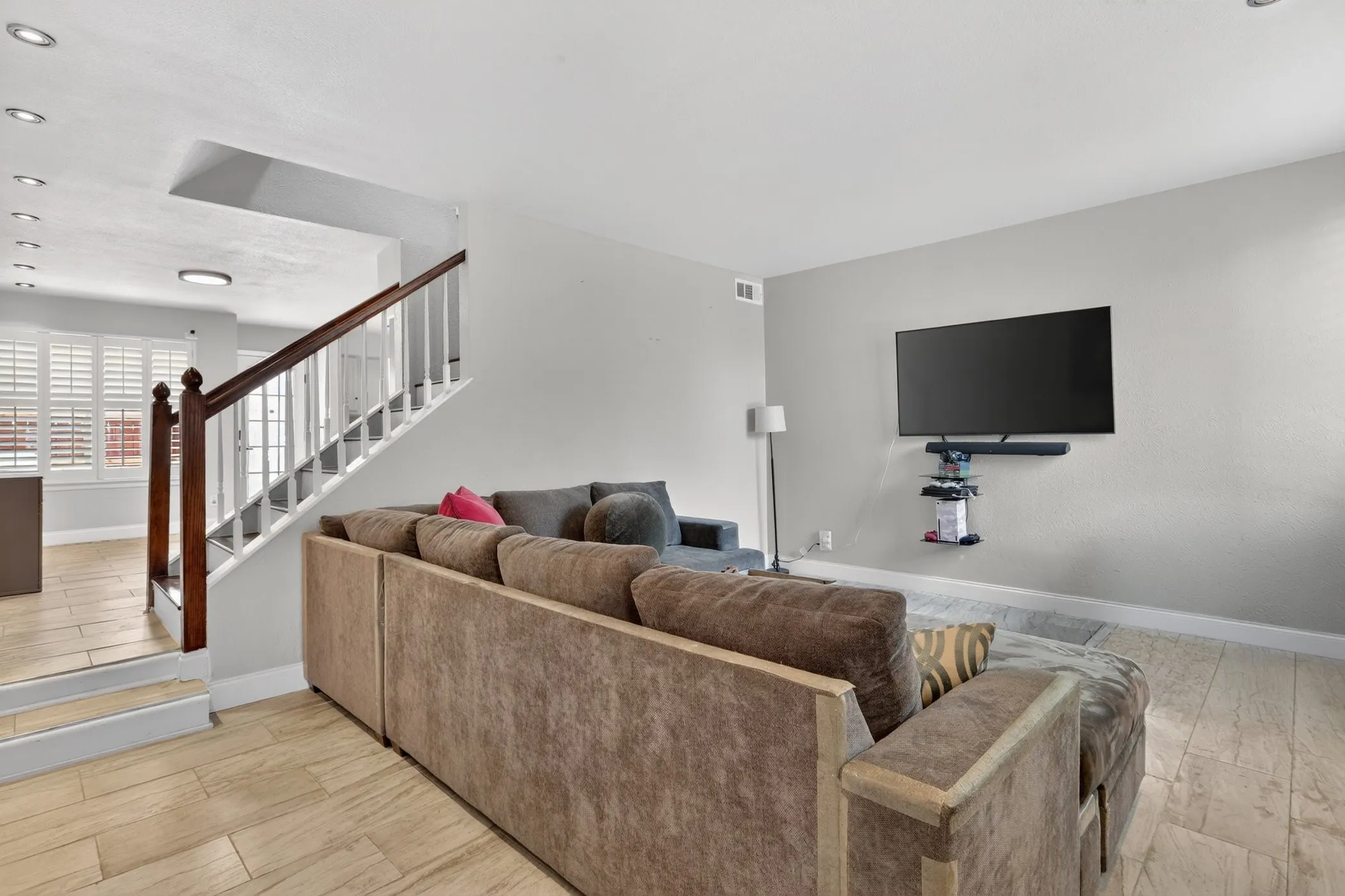 Living area featuring light wood-finish flooring, a contemporary staircase with white balusters and a dark wood handrail, recessed ceiling lighting, and a wall-mounted entertainment setup