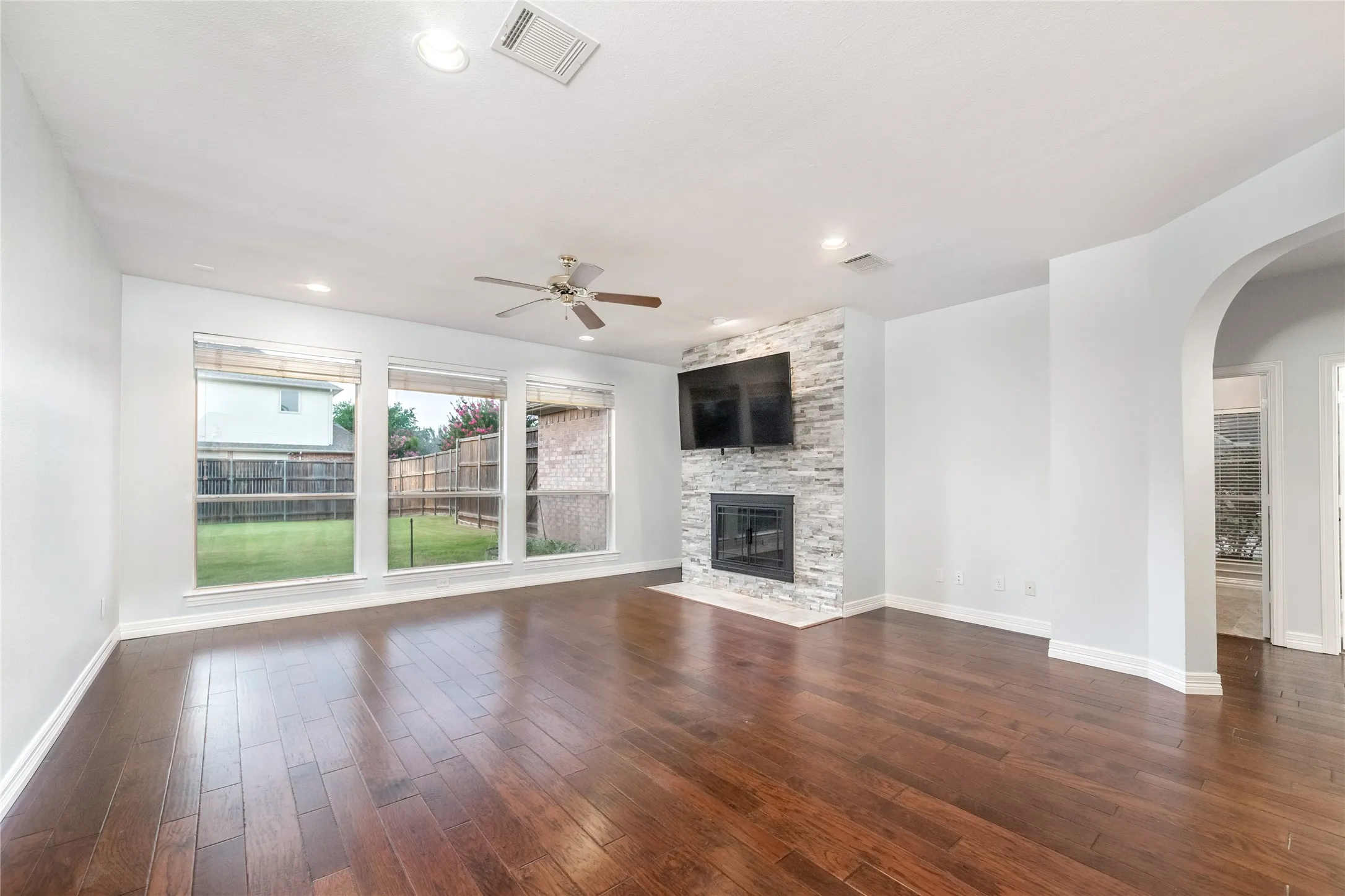 Spacious living area featuring wood-finish flooring, a stone-clad fireplace, a ceiling fan with integrated lighting, recessed lighting, and large windows overlooking the backyard