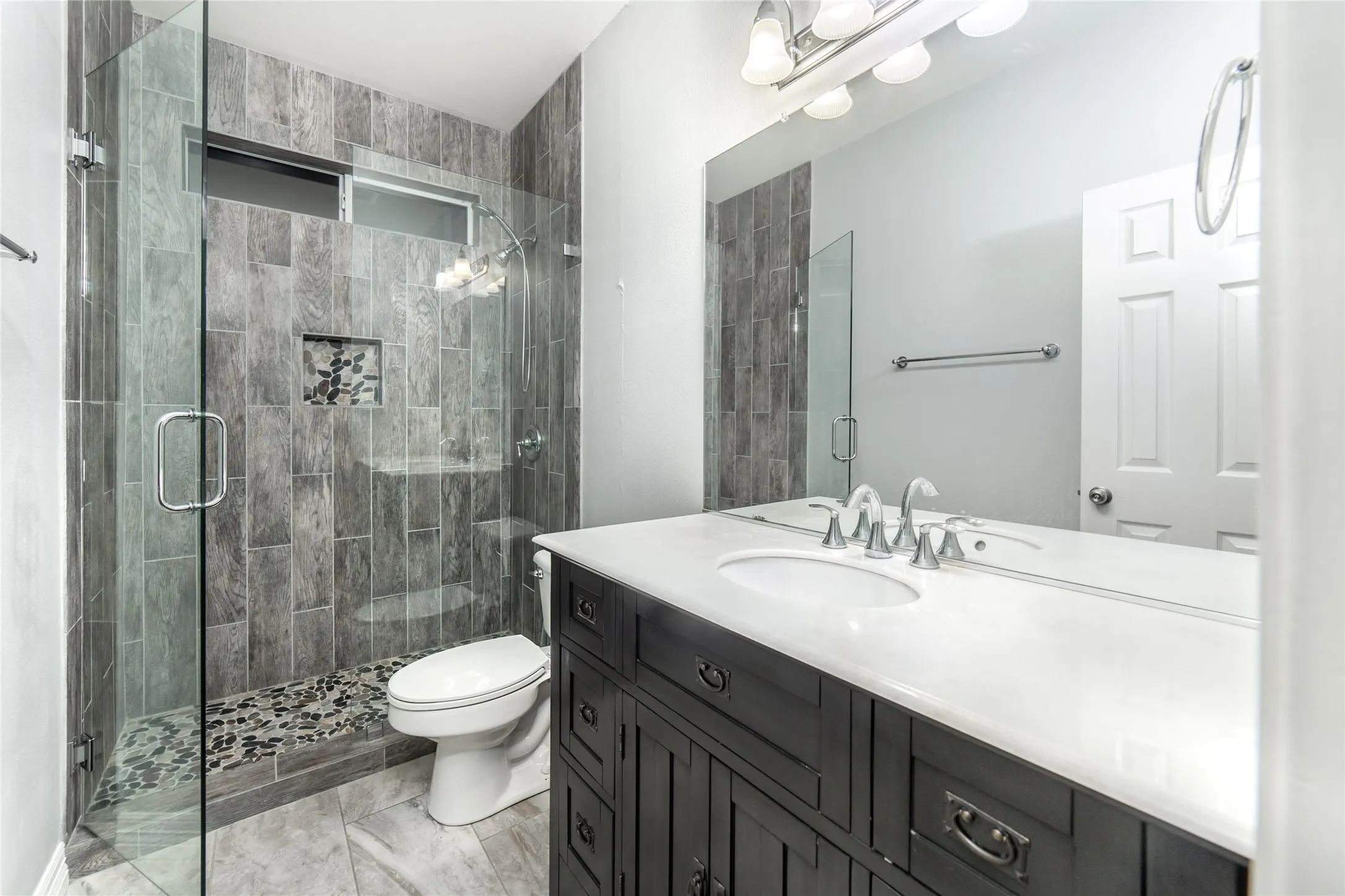 Modern bathroom with a glass-enclosed shower featuring gray wood-finish wall tile, a mosaic stone shower floor, and a built-in shower niche