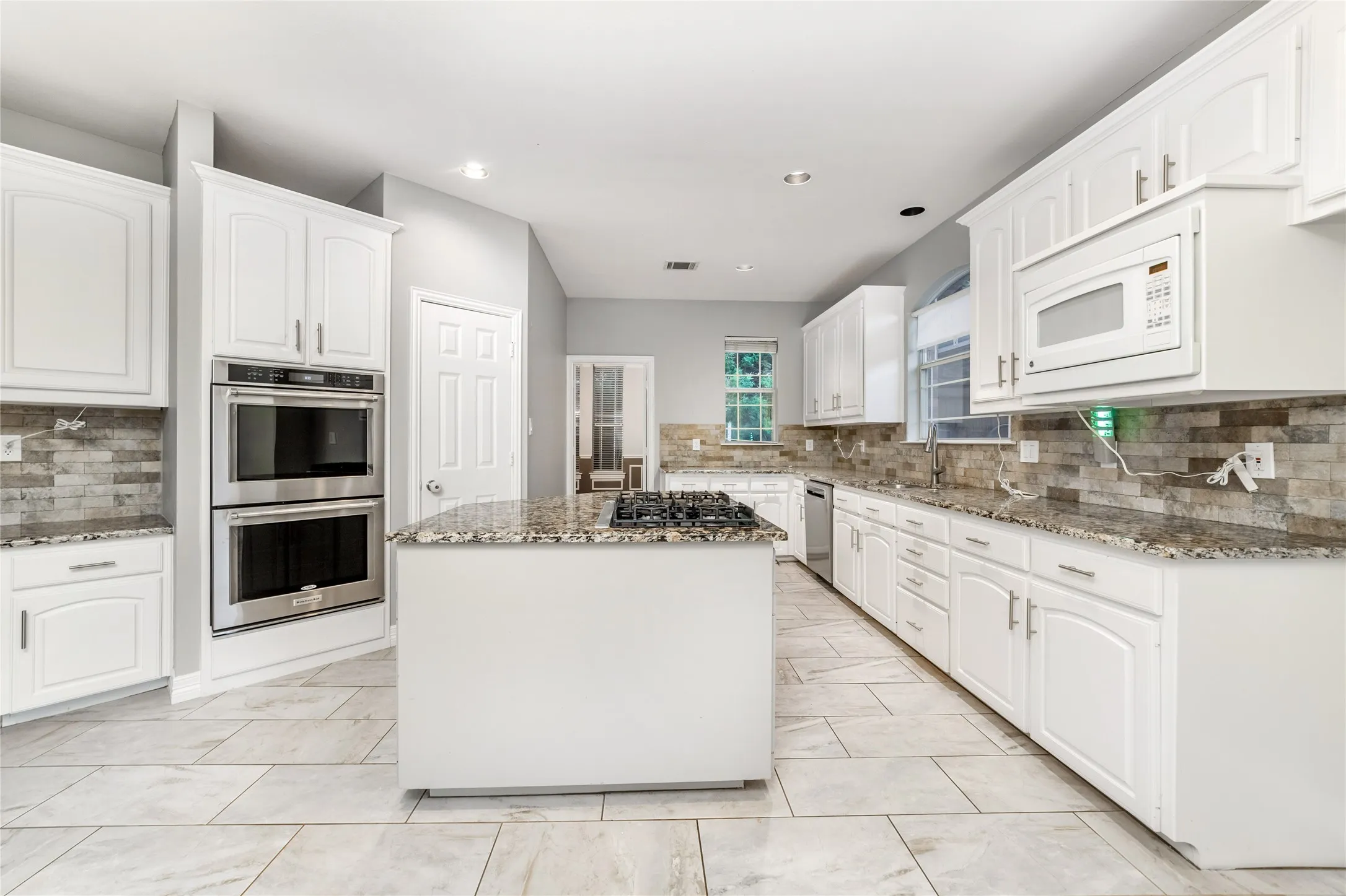 Spacious kitchen featuring an island with a gas cooktop, granite countertops, tile backsplash, white cabinetry, and stainless steel double ovens