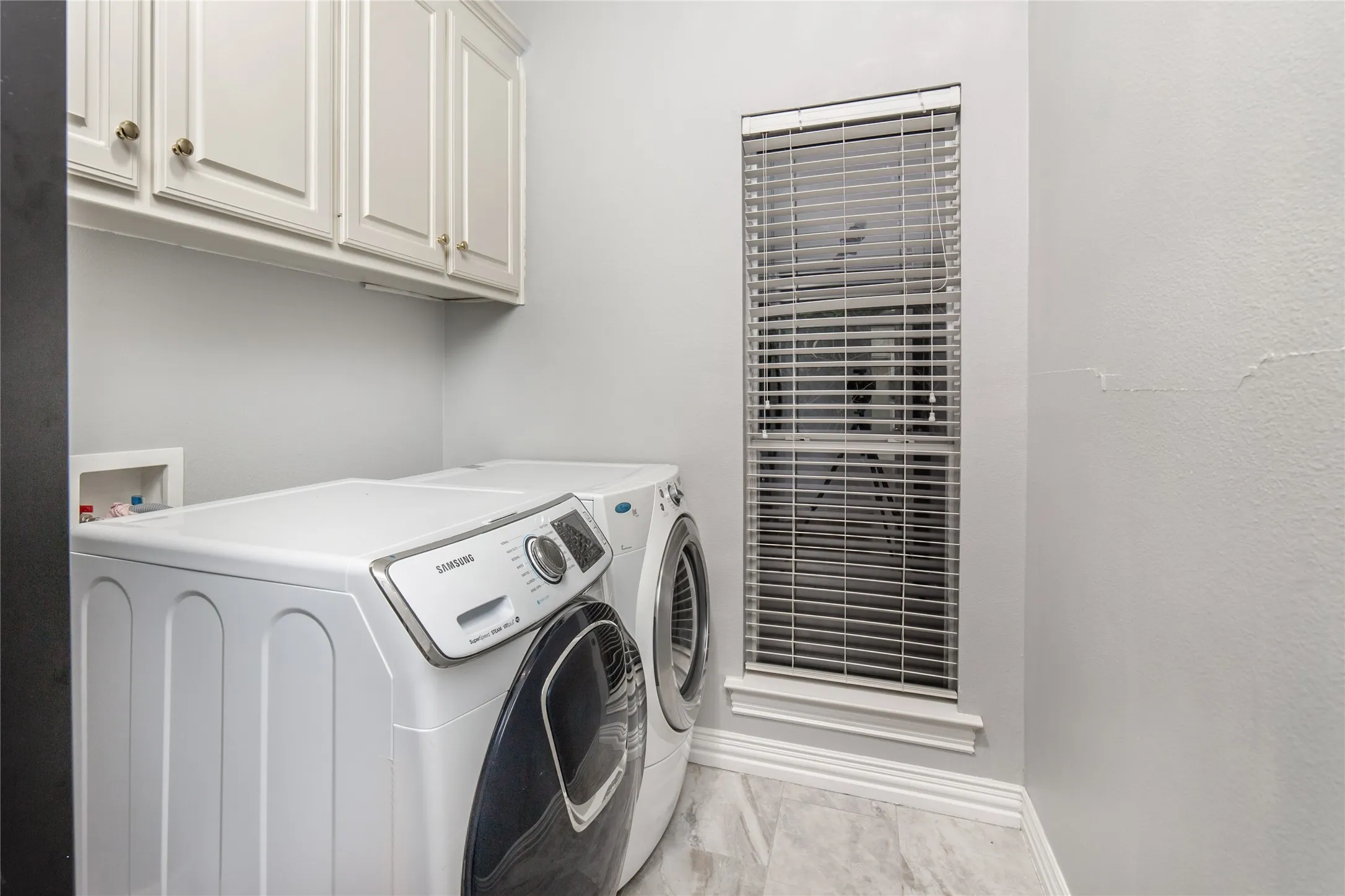 Dedicated utility space featuring overhead white cabinetry, a single window with blinds, and light-toned tile flooring
