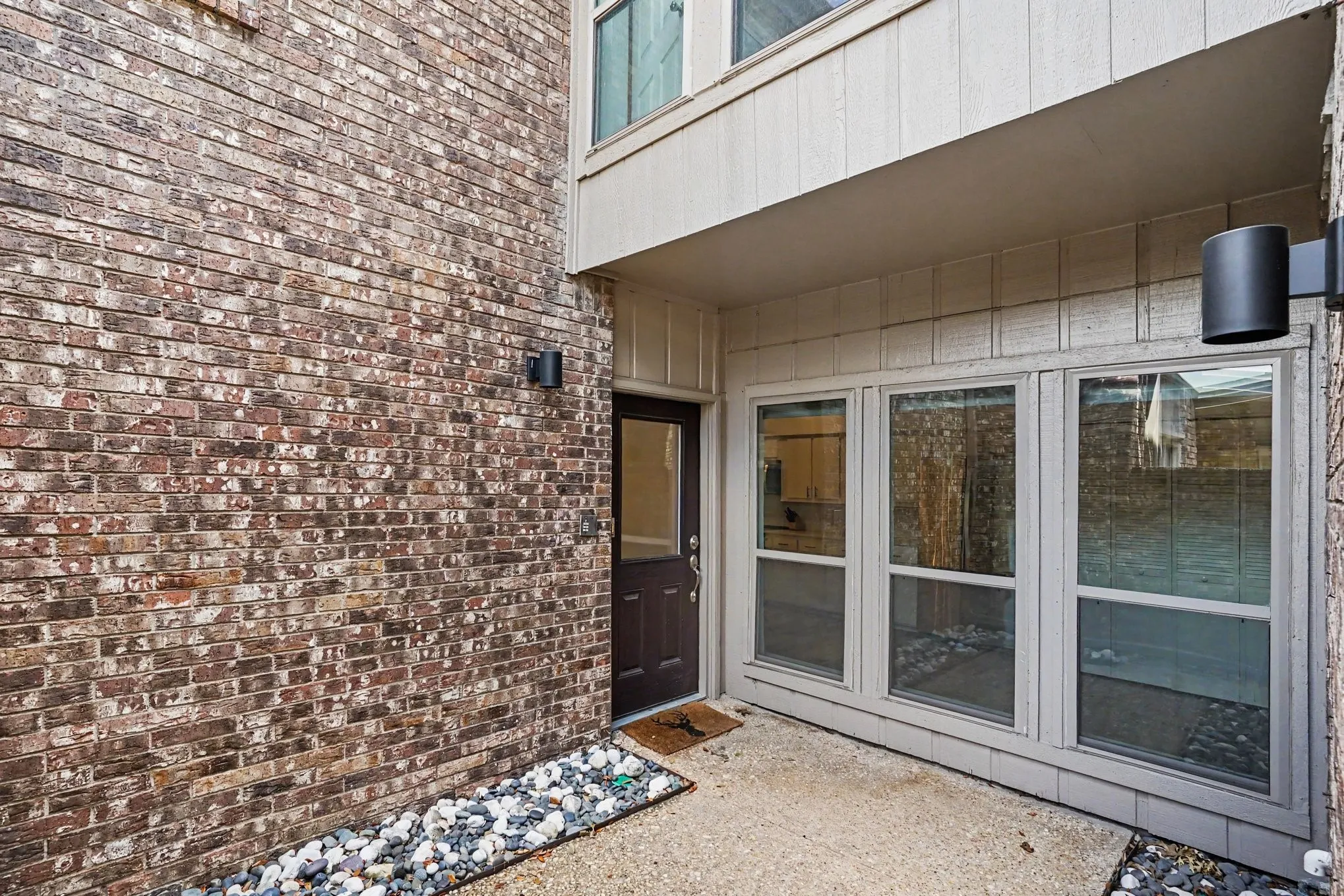 Exterior patio area featuring a textured concrete surface, a dark paneled door, three large windows, and a brick facade