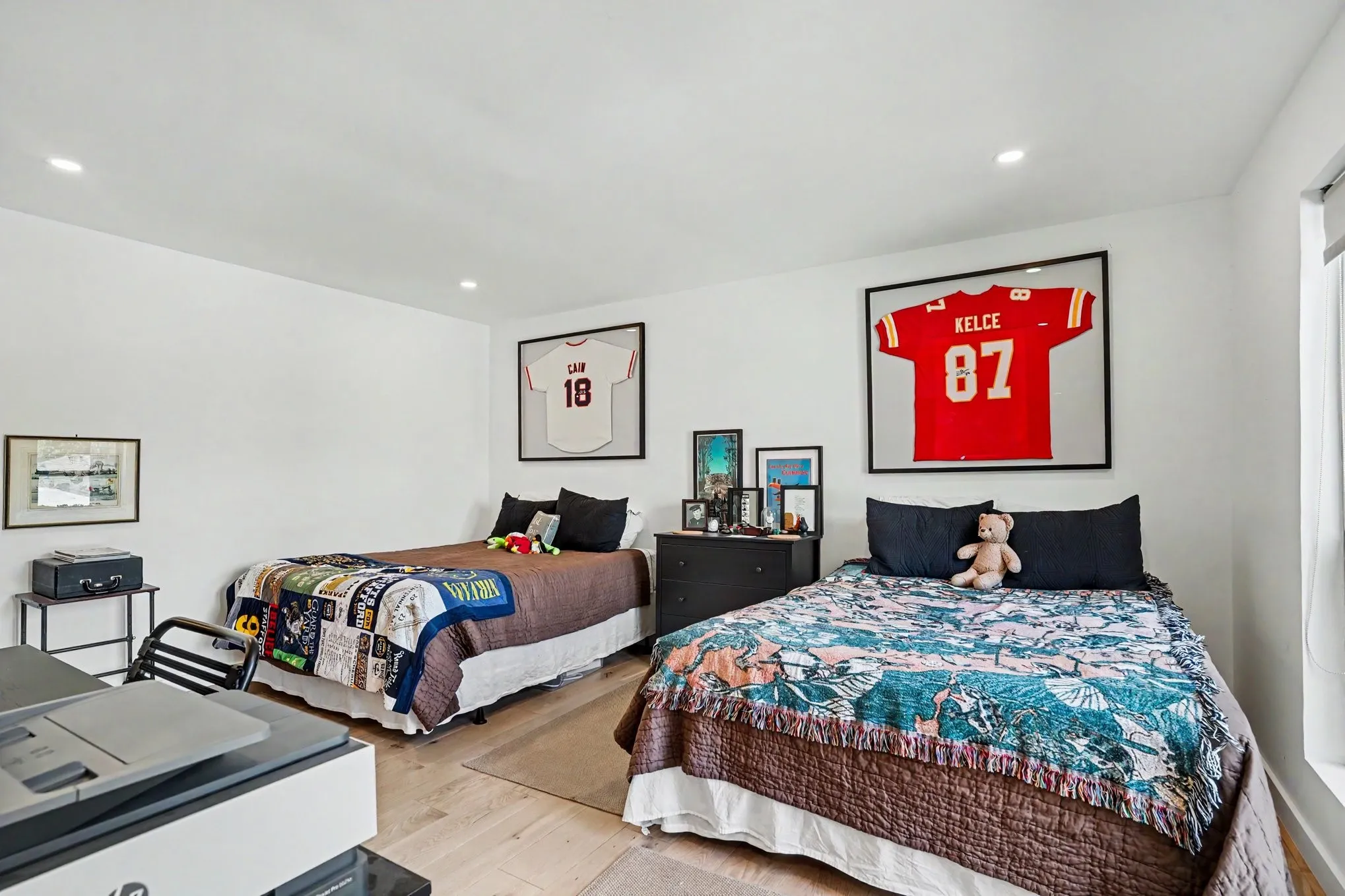 Bright room featuring two beds, white walls, wood-finish flooring, recessed lighting, and a window with a roller shade
