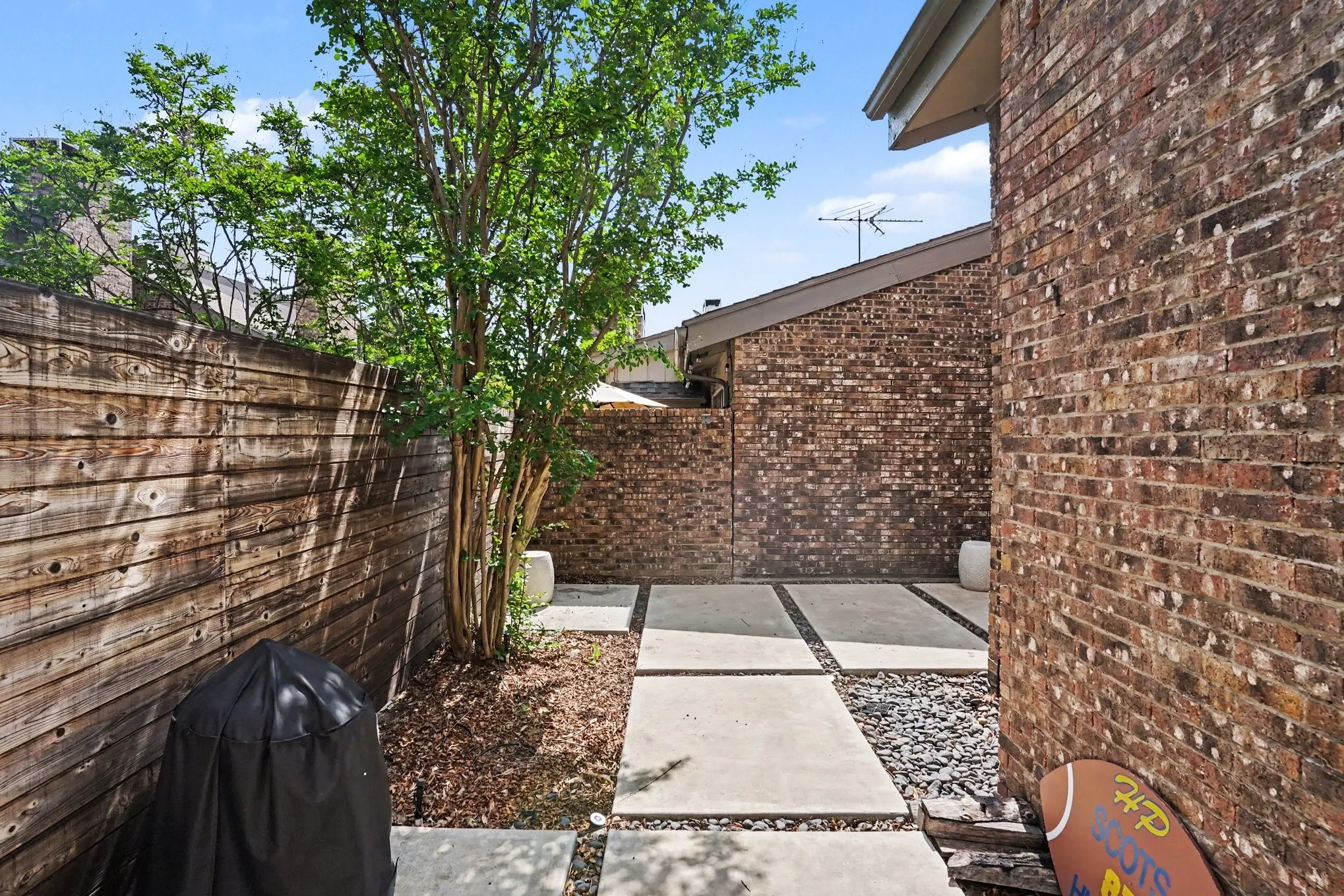 Private outdoor space featuring a wood fence, mature tree, brick exterior, and concrete paver walkway with dark gravel accents