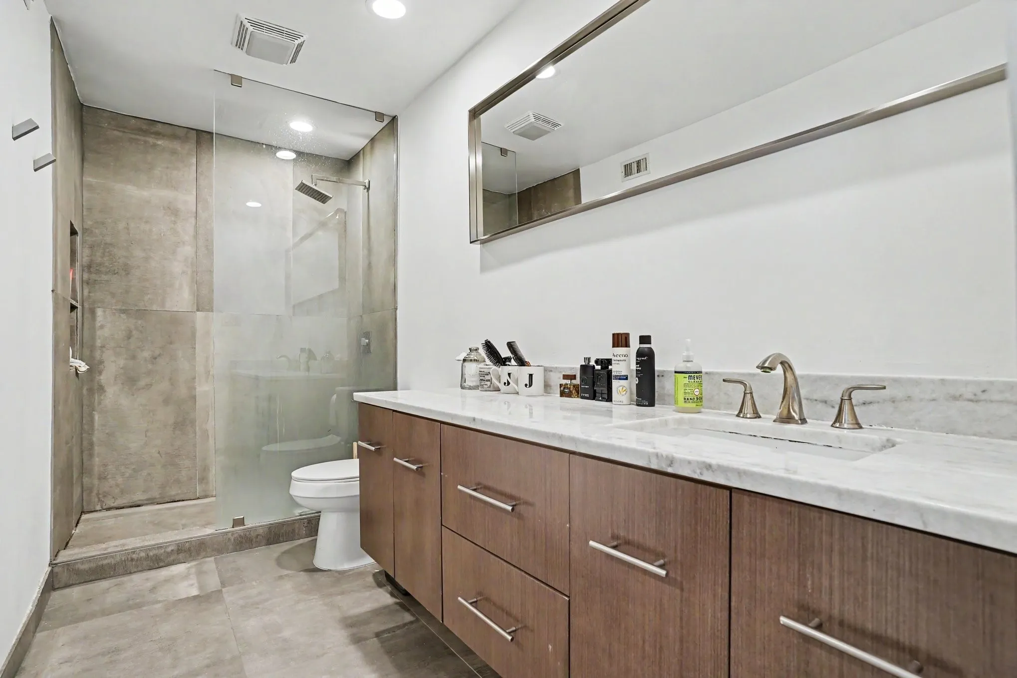 Bathroom featuring a frameless glass shower enclosure, a dual-sink vanity with a light-toned countertop, wood-finish cabinetry, and brushed nickel fixtures