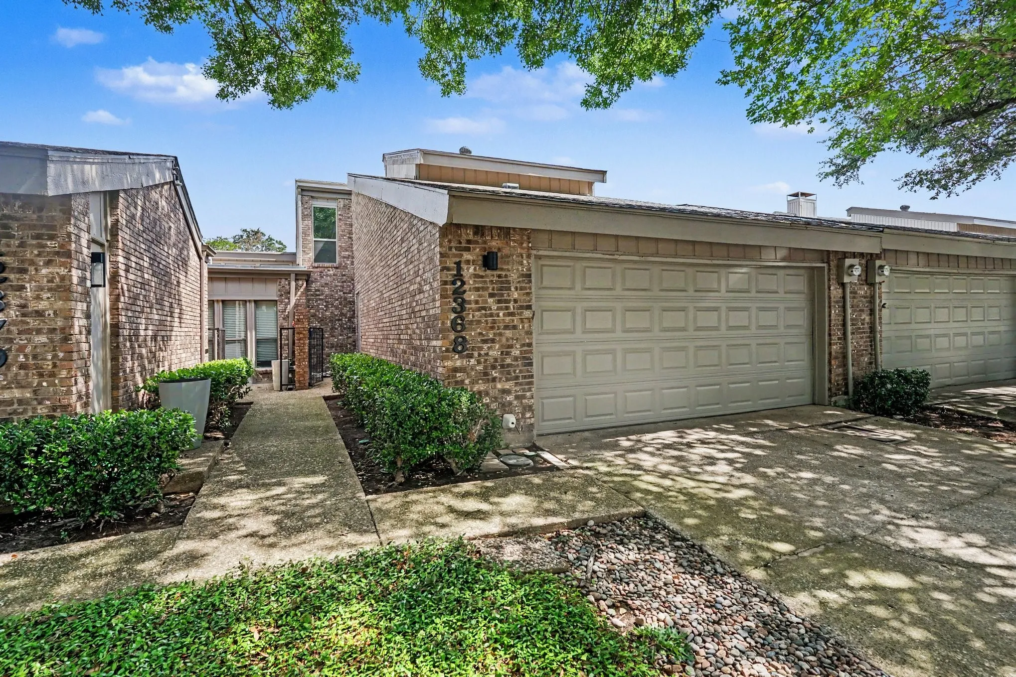 Brick exterior residence featuring two attached garages with panel doors, a concrete walkway, mature shrubbery, and integrated exterior lighting fixtures
