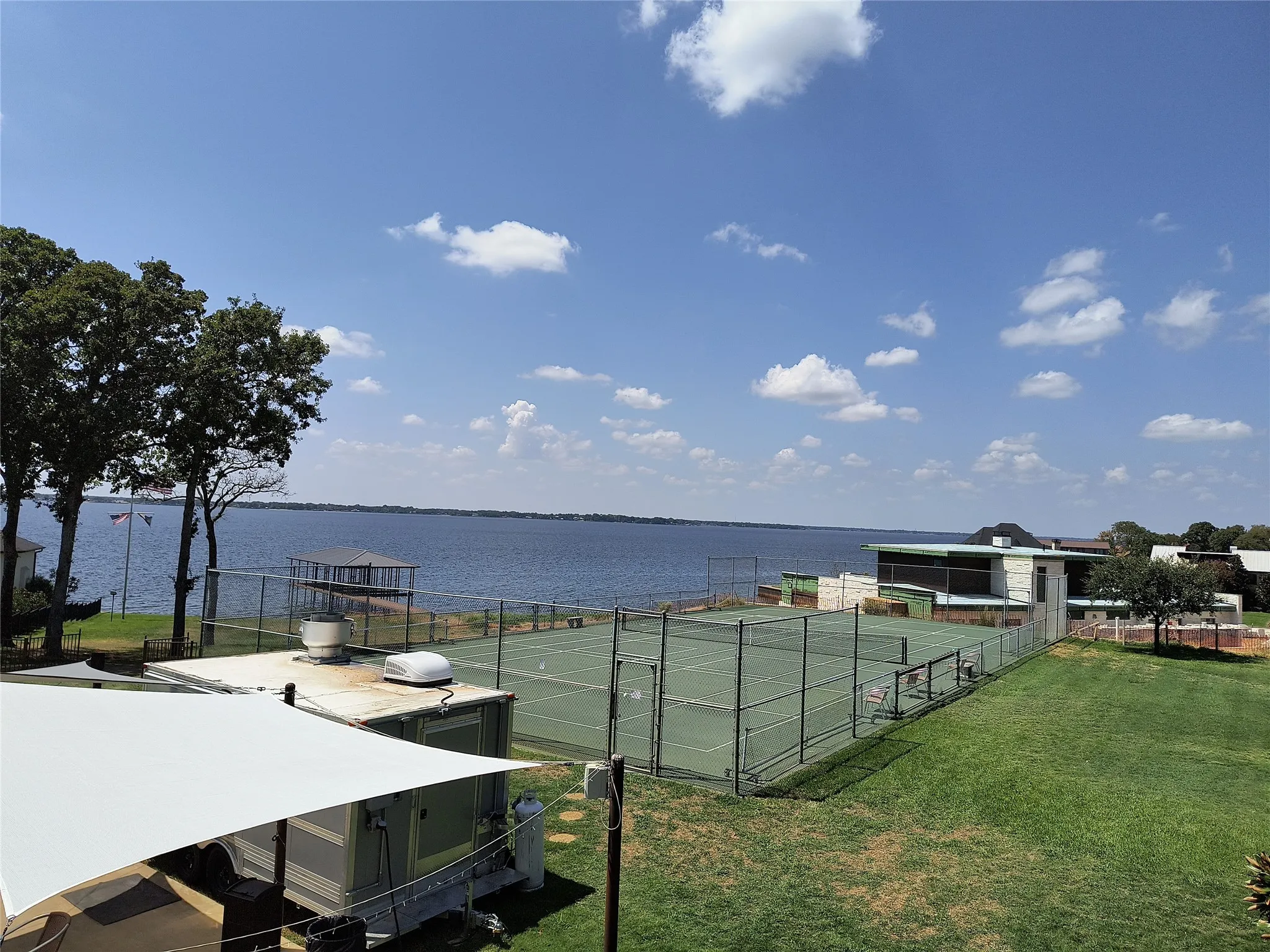 Lakeside property featuring a full-size tennis court with perimeter fencing, expansive green lawn, and mature trees