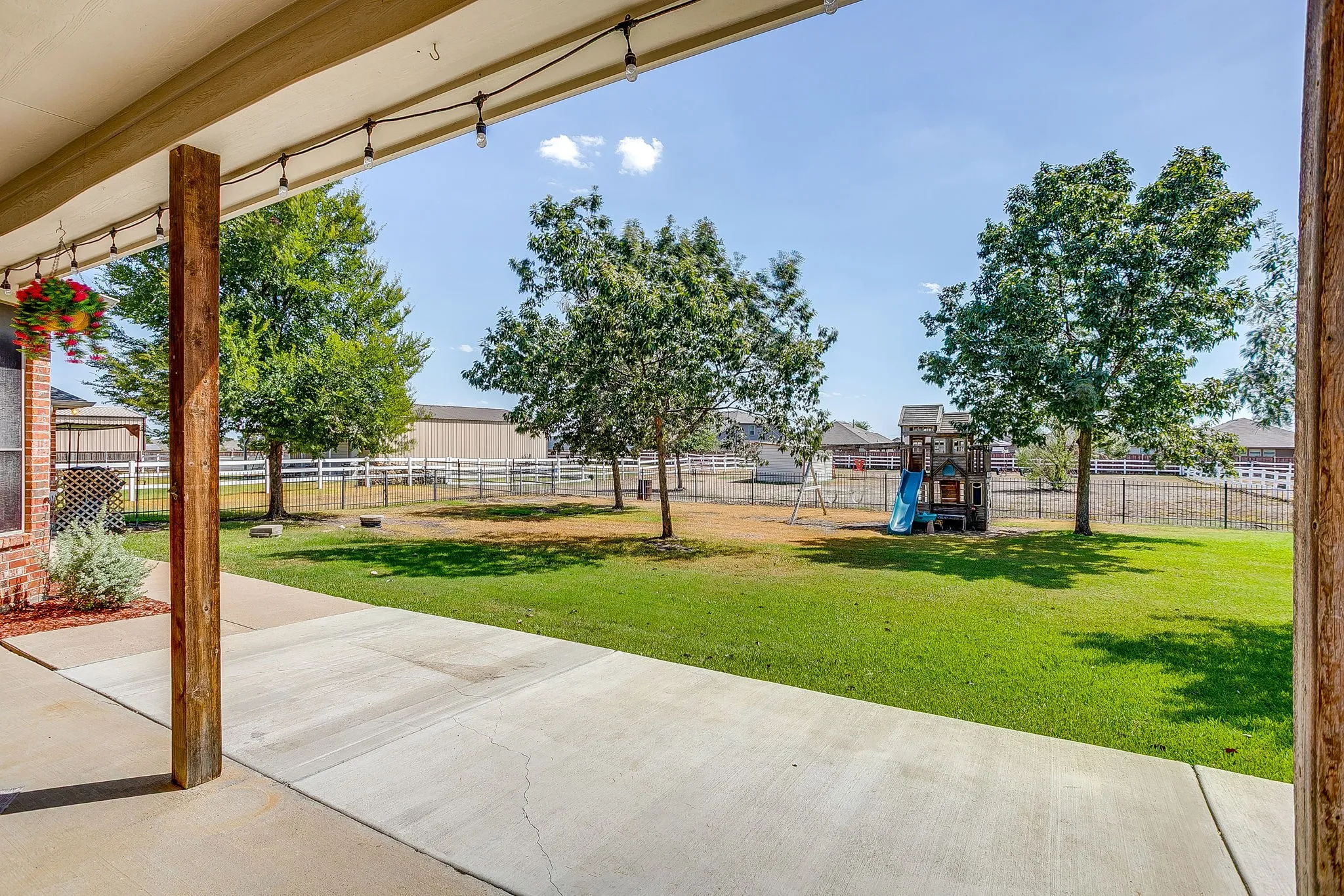 Covered patio with string lighting, overlooking a green lawn with mature trees and a fenced perimeter