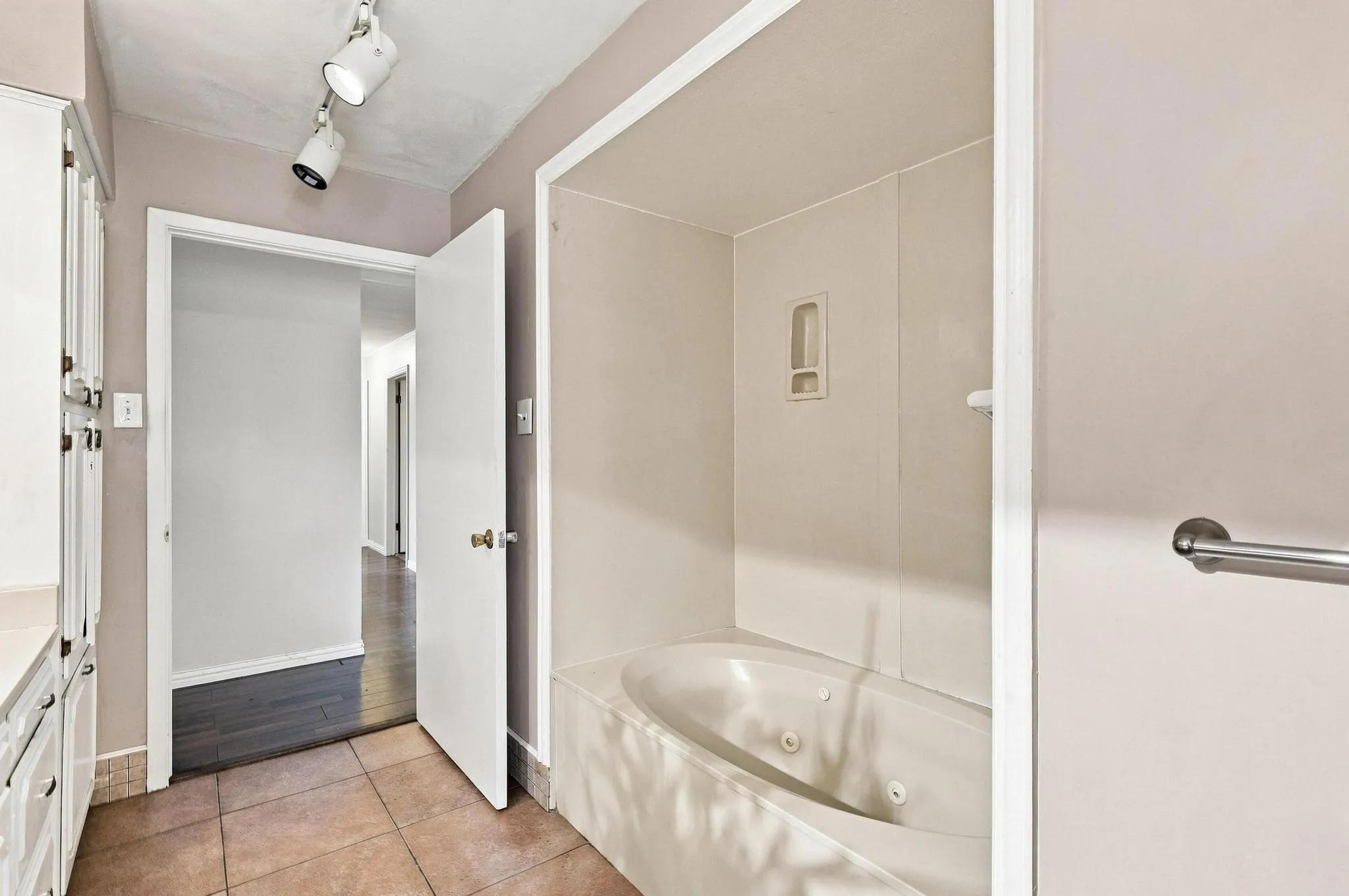 Jetted soaking tub with built-in storage niche, track lighting, white cabinetry with integrated countertop, and brown tile flooring