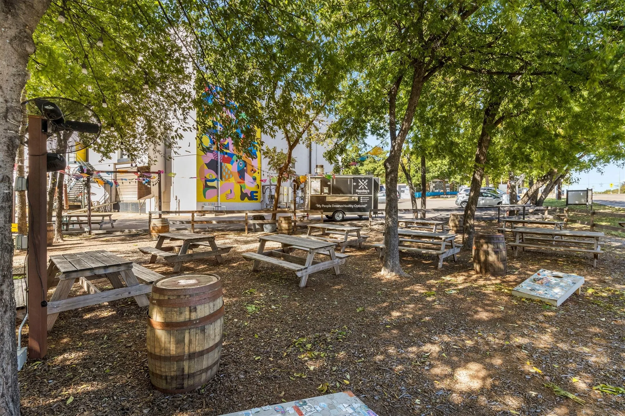 Outdoor gathering space featuring multiple picnic tables, mature trees, and a gravel ground cover