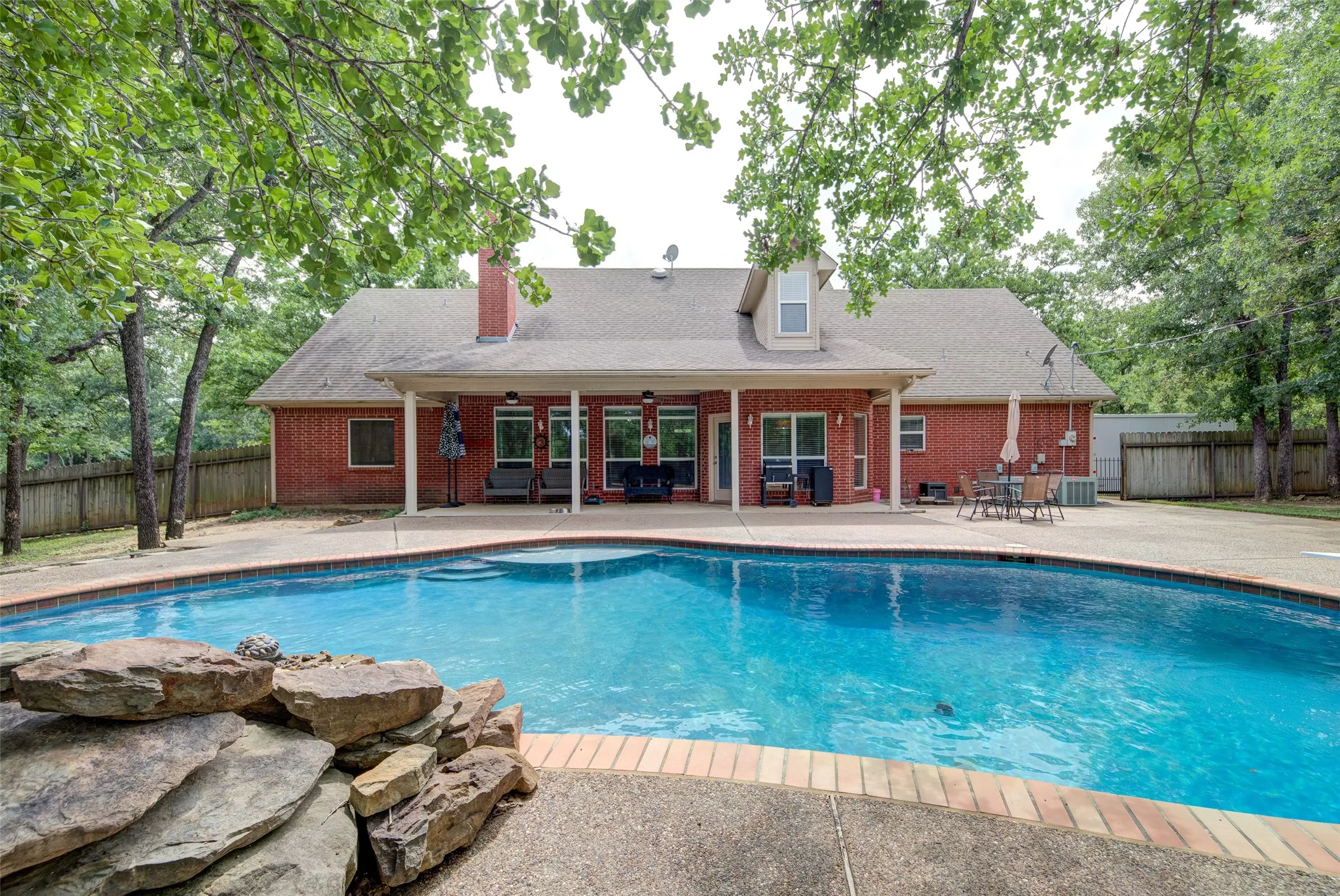 Expansive backyard featuring a large swimming pool with stone accents, a red brick exterior, a covered patio, and mature trees providing natural canopy