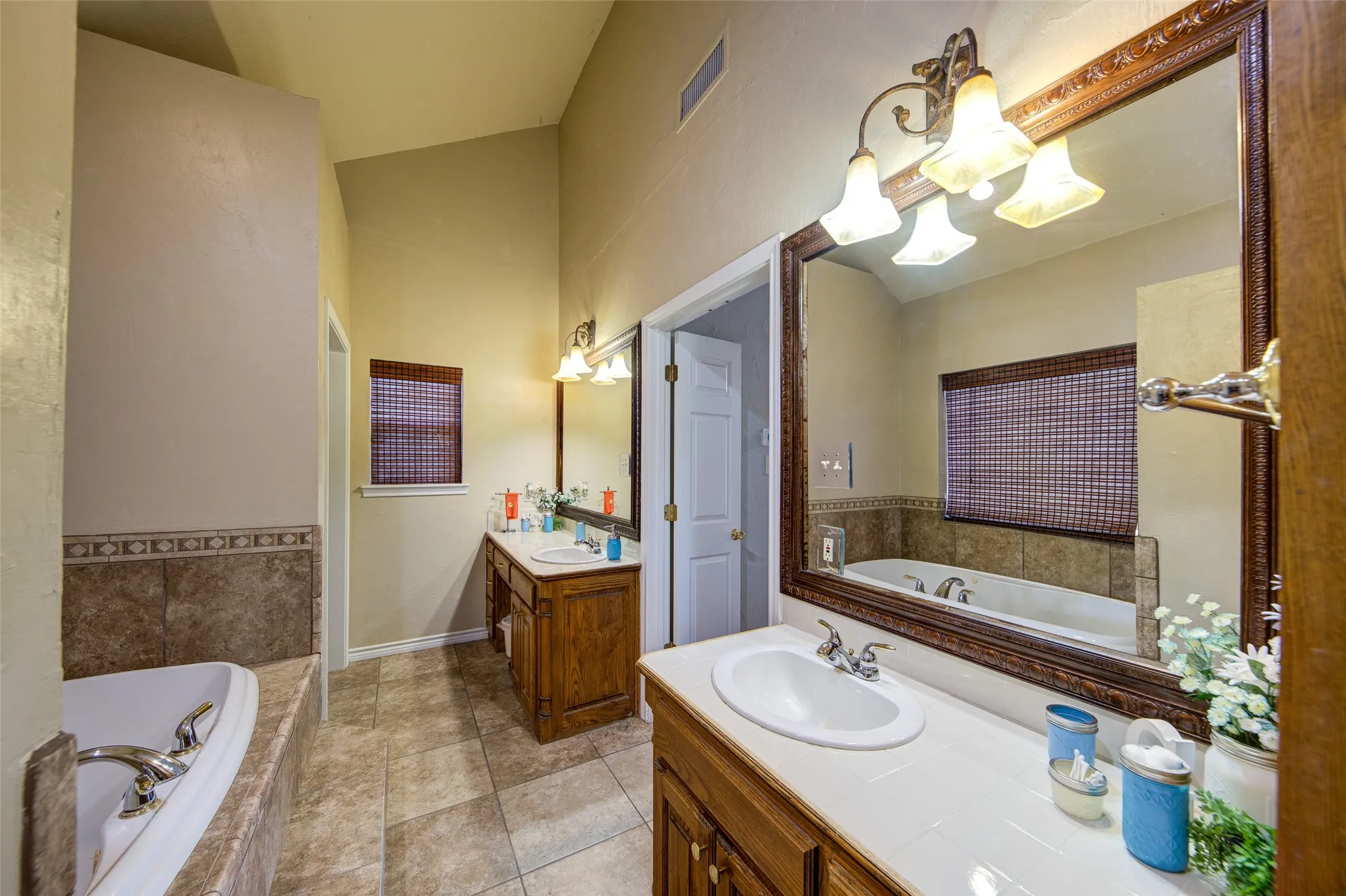 Bathroom featuring dual wood vanities with integrated sinks, a large soaking tub with tiled surround, patterned tile flooring, and vaulted ceilings