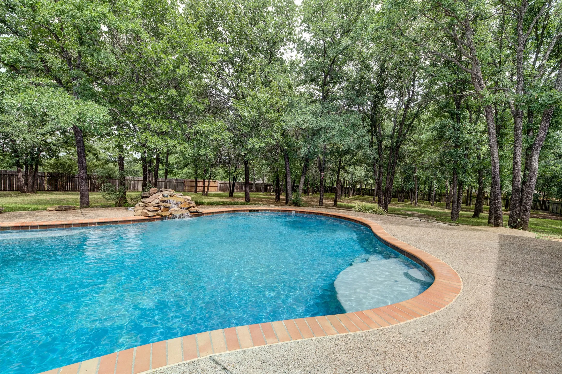 Curved inground pool featuring a rock water feature, integrated entry steps, brick coping, and a surrounding aggregate patio