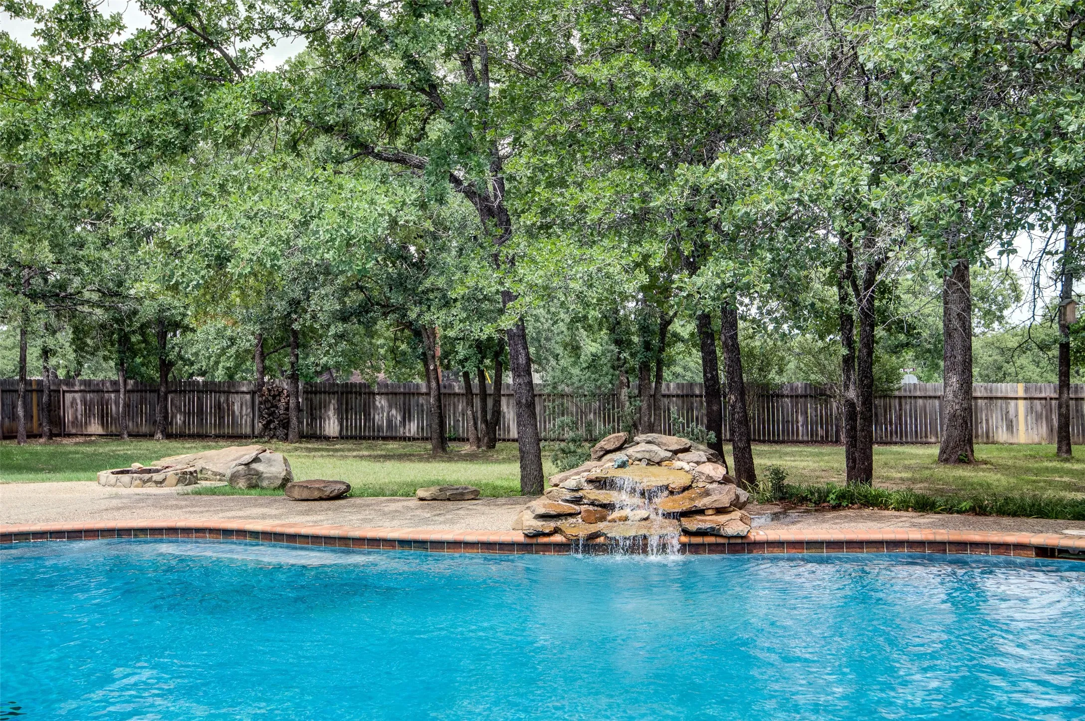 Outdoor swimming pool featuring a rock waterfall, surrounding concrete decking with tile coping, and a grassy backyard with mature trees