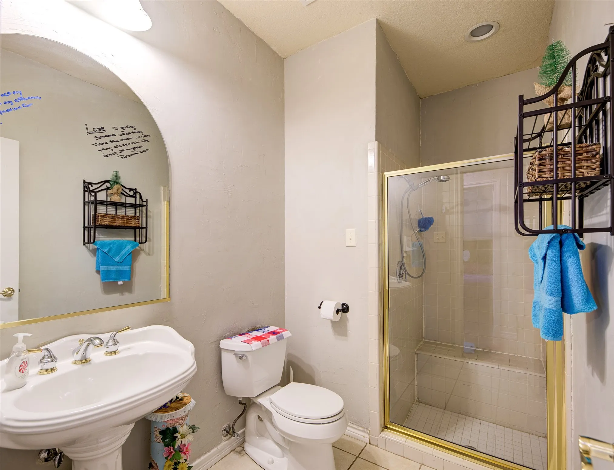 Pedestal sink with brass-finish dual-handle faucet, glass-enclosed shower with gold-tone frame, recessed lighting, and wall-mounted shelving