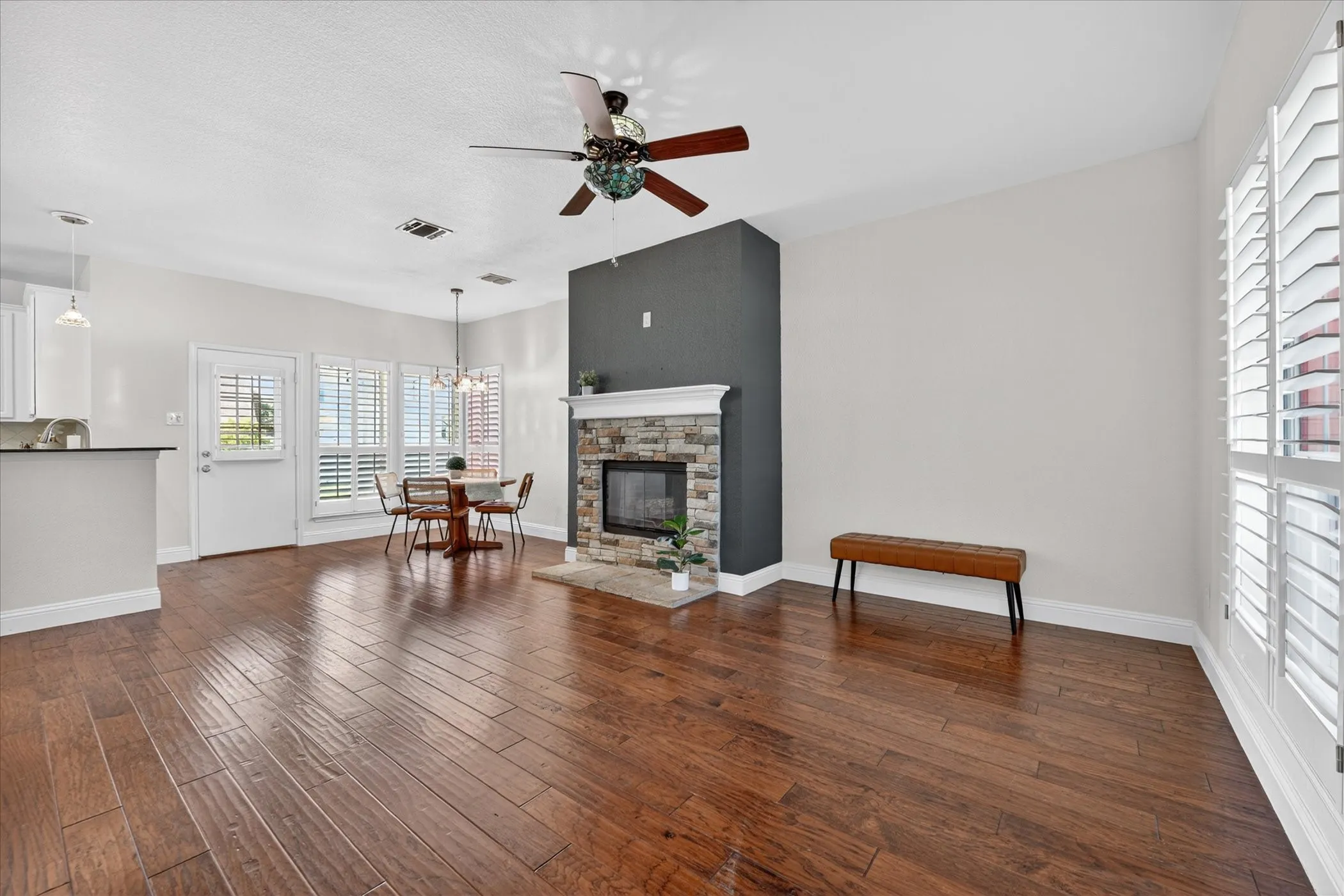 Spacious room featuring wood-finish flooring, a stone fireplace with a dark accent wall, and multiple windows with plantation shutters
