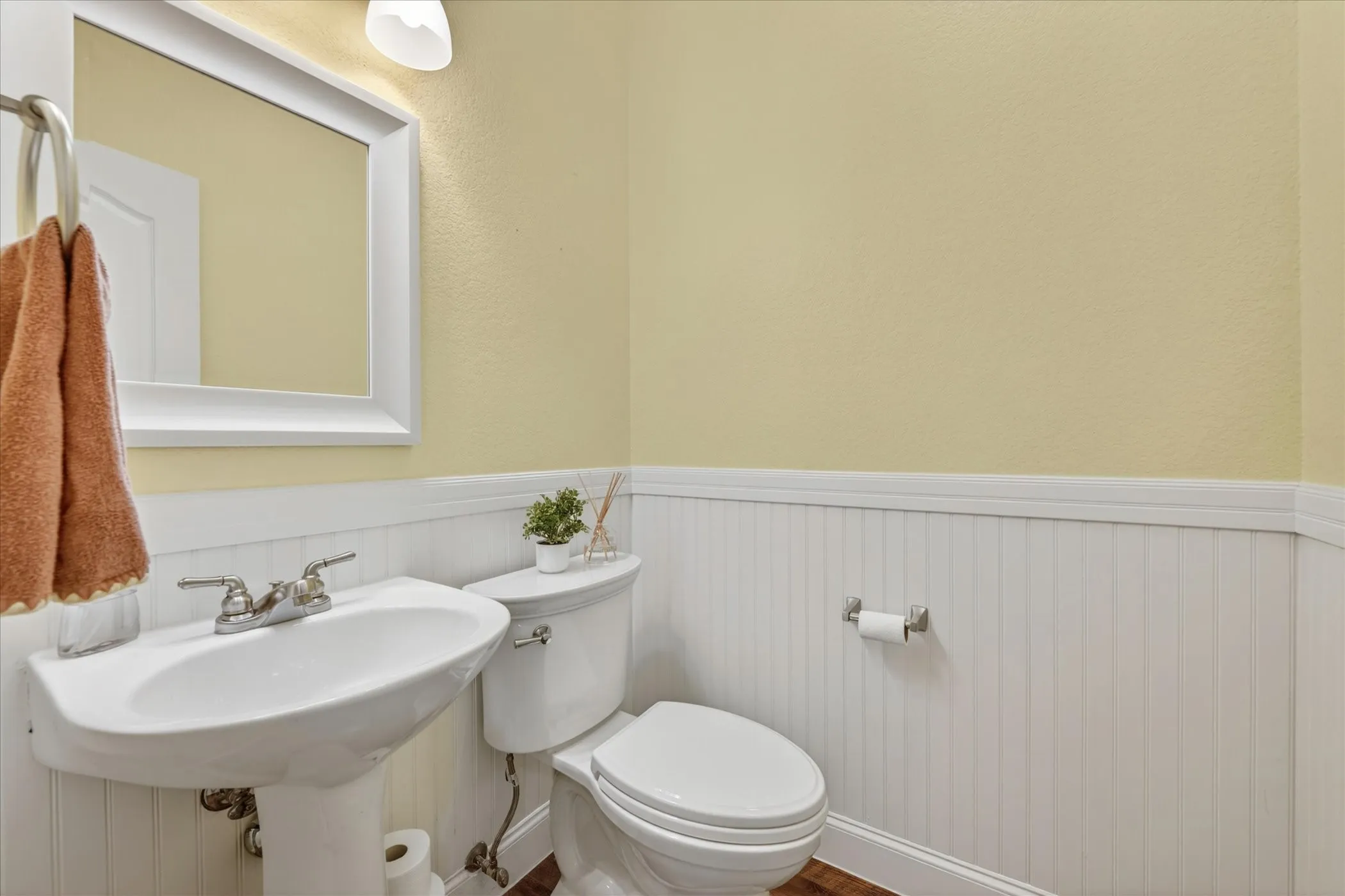 Pedestal sink with chrome faucet, white beadboard wainscoting, light yellow walls, framed wall mirror, and a white toilet