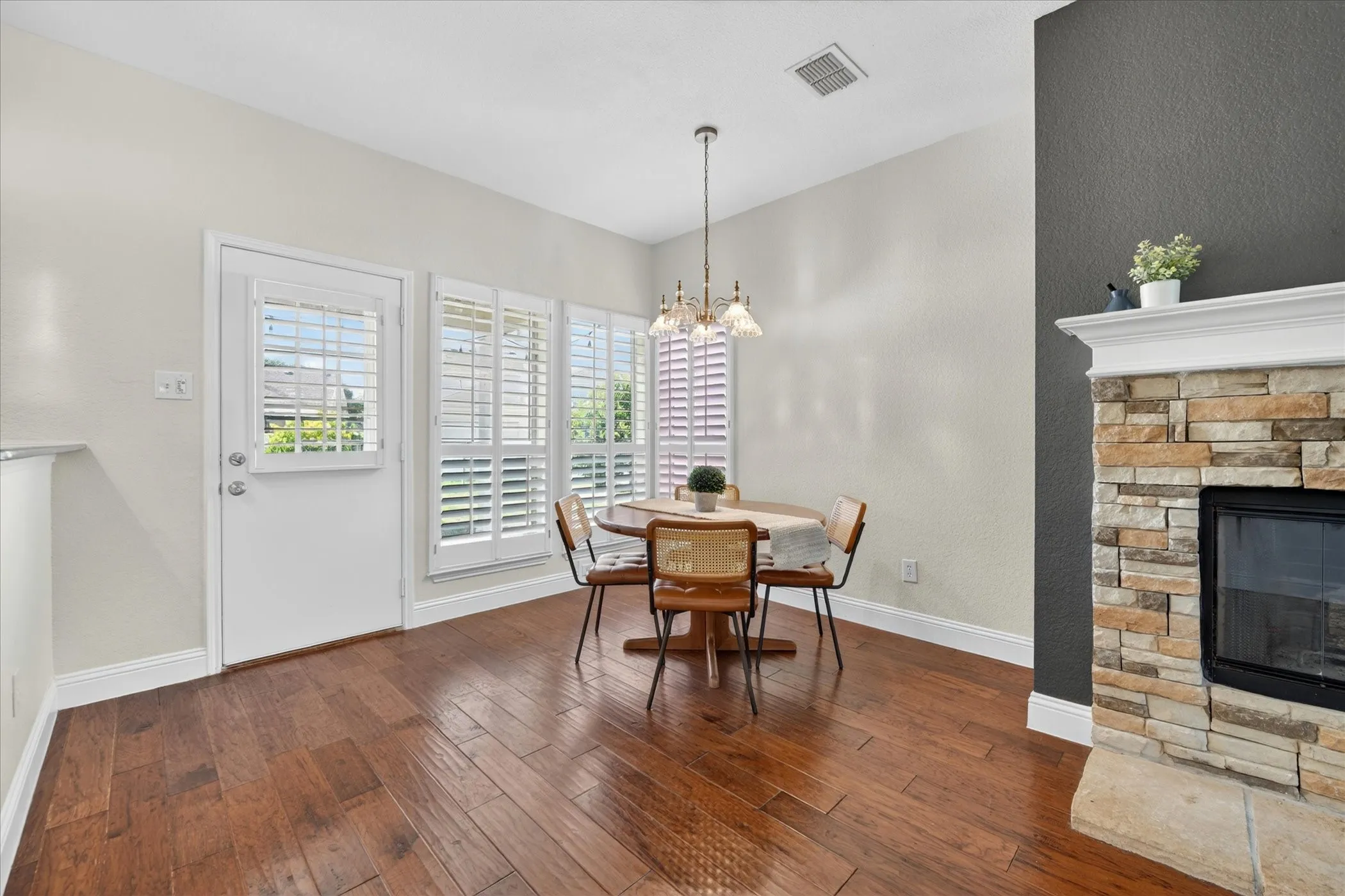 Dining area featuring wood-finish flooring, a stone-clad fireplace with a white mantel, and a door with glass panels and plantation shutters