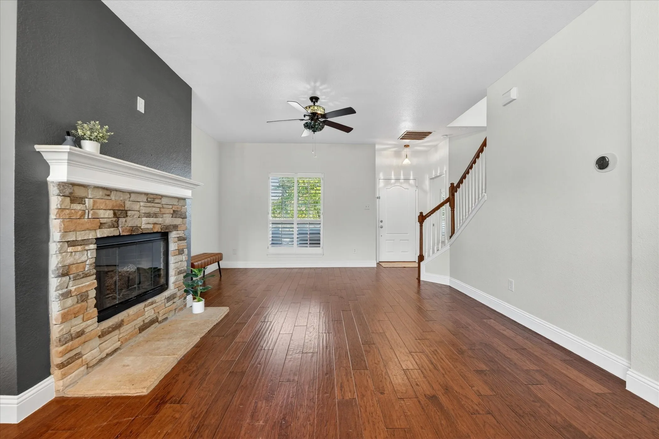 Spacious living area featuring wood-finish flooring, a stone-faced fireplace with a white mantel, and a ceiling fan