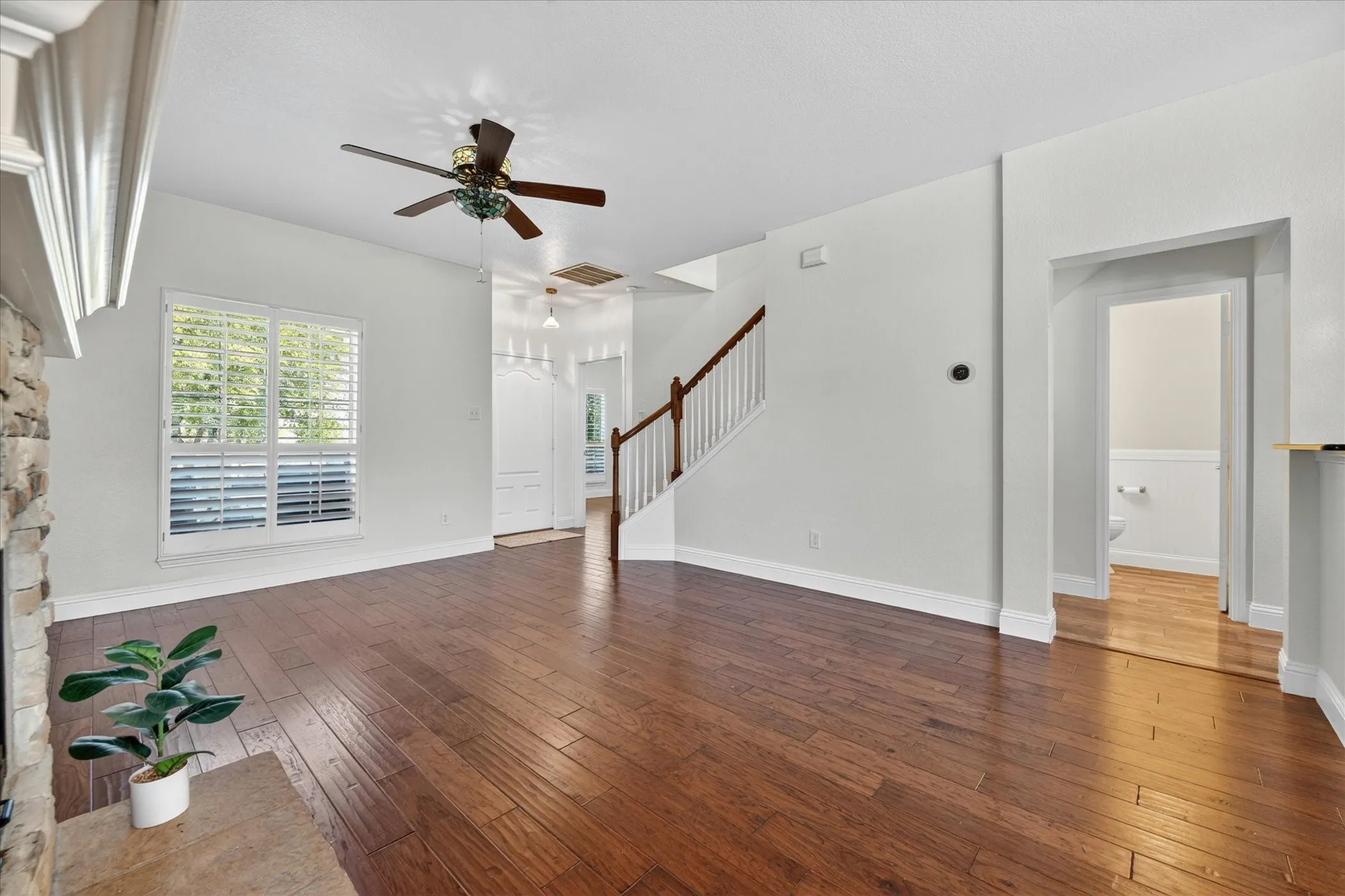 Spacious living area featuring wood-finish flooring, a stone-faced fireplace with a mantel, a ceiling fan, window shutters, and a visible staircase with wood newel posts and handrail