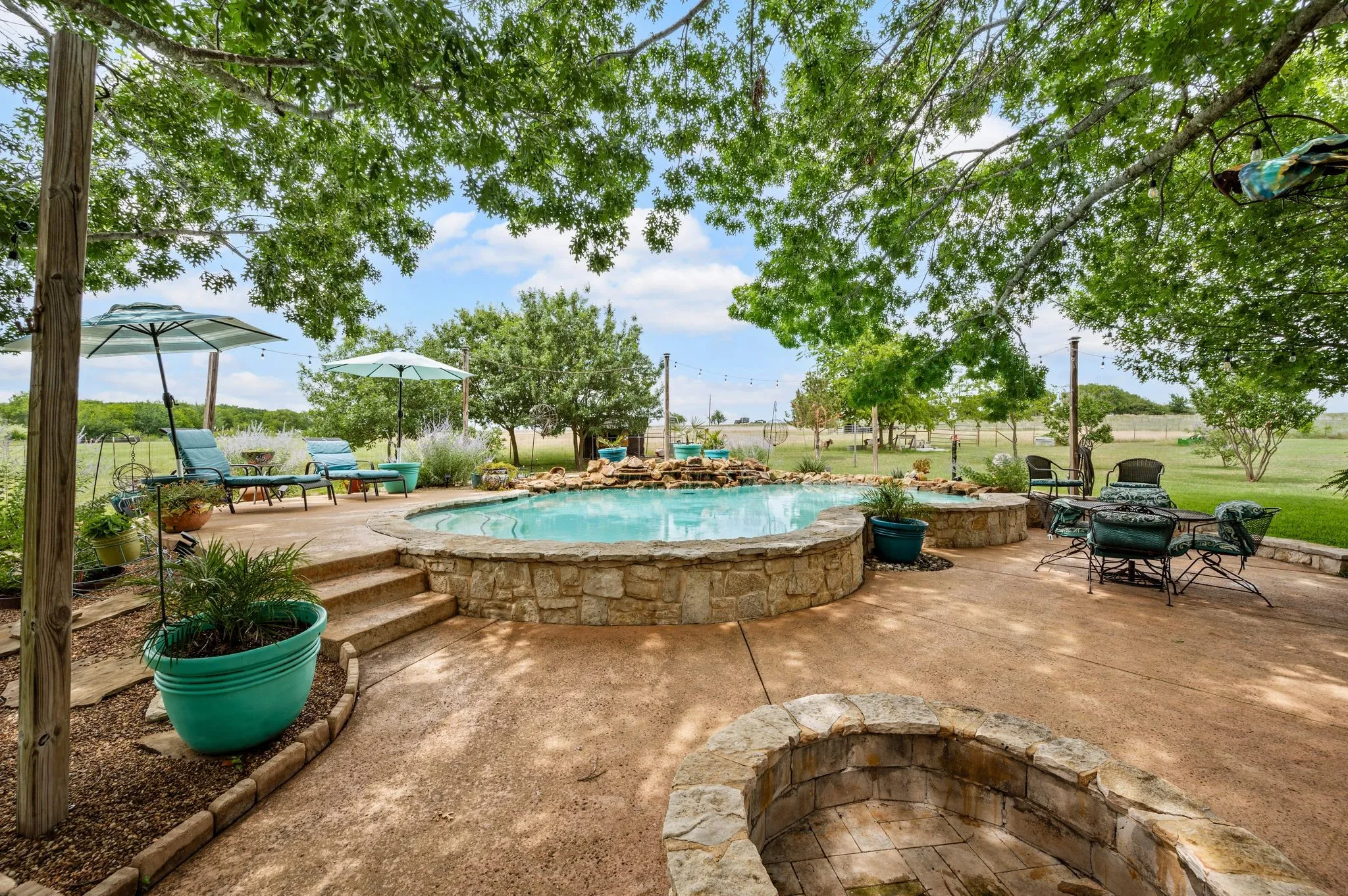 Outdoor living area featuring a gunite pool with a waterfall, a built-in fire pit, multiple seating areas on the patio, and mature trees providing natural shade