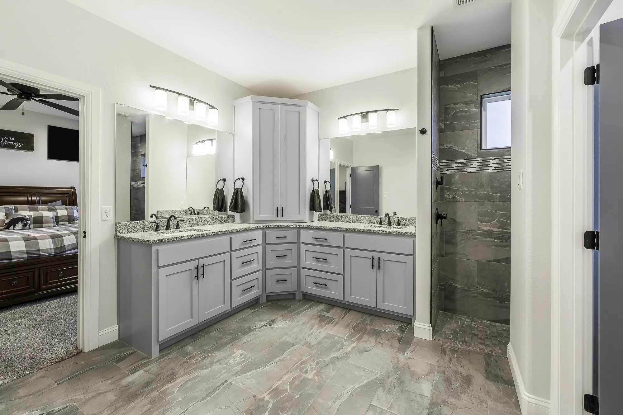 Spacious bathroom featuring marble-look tile flooring, dual vanities with light gray cabinetry and granite countertops, and a walk-in shower with dark gray tile and accent banding