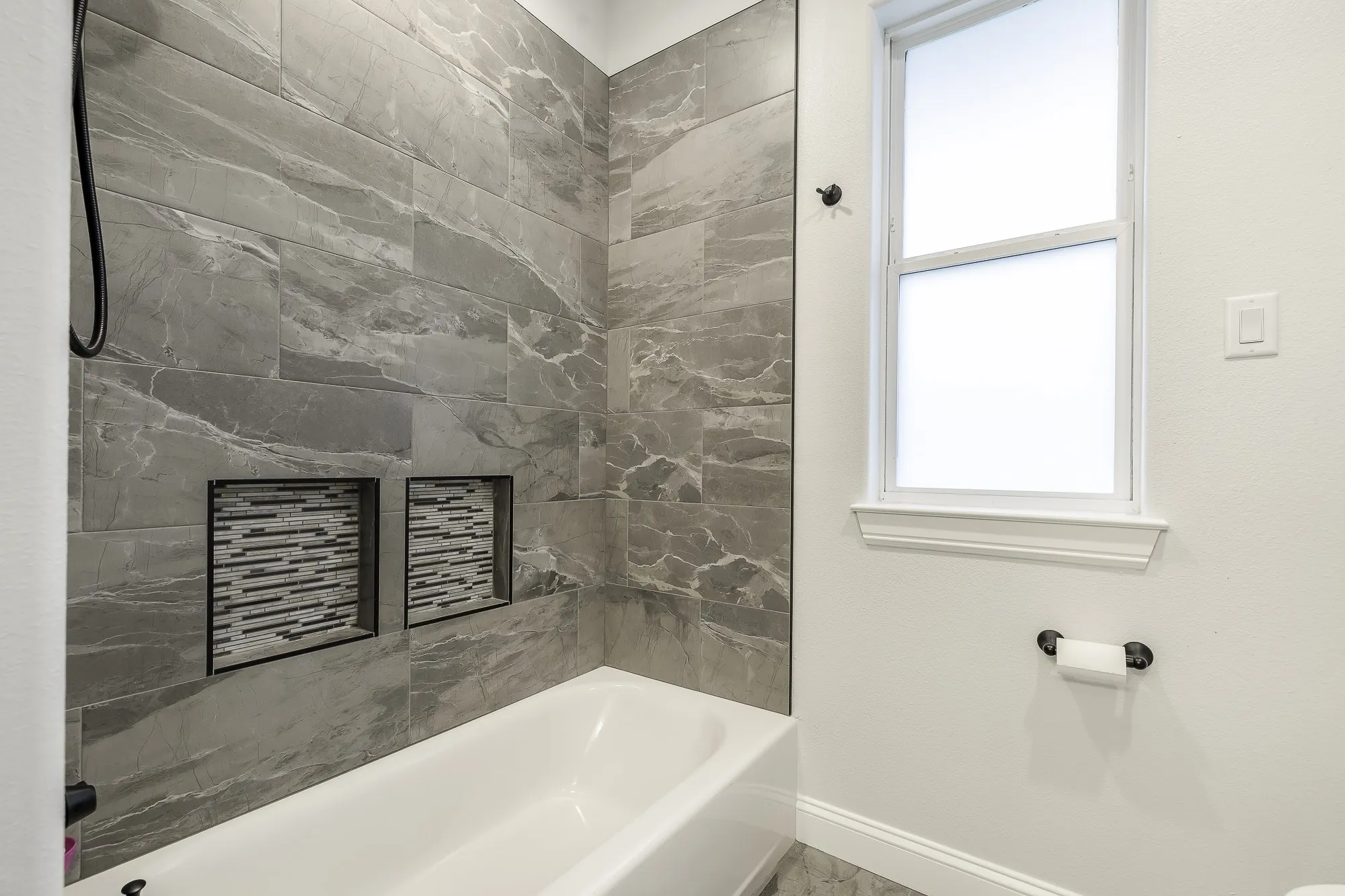 Bathtub and shower combination featuring large format grey marbled wall tiles and dual recessed niches with mosaic tile backings